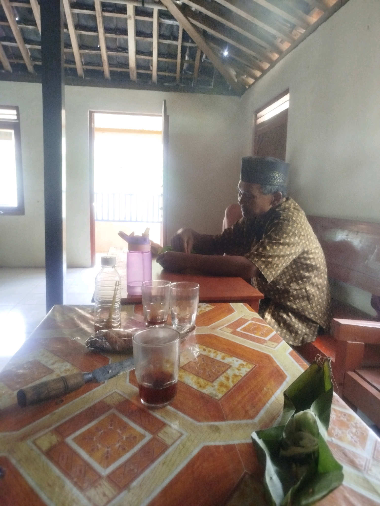 An indoor scene in a rustic Indonesian dwelling in Ponjong, Indonesia, features a man wearing a black peci hat and a patterned brown shirt seated on a wooden bench. He is looking to his left, with his arm resting on a wooden surface. In the foreground, a table is set with several items. A patterned tablecloth covers the table, on which sit three glasses, one containing a dark liquid, a water bottle, and a purple shaker bottle. A knife with a wooden handle is also present on the table. To the right of the frame, a portion of a banana leaf parcel containing food is visible. The room has an unfinished wooden ceiling with exposed beams and light entering from doorways and windows, illuminating the space. The overall impression is of a humble, home environment.