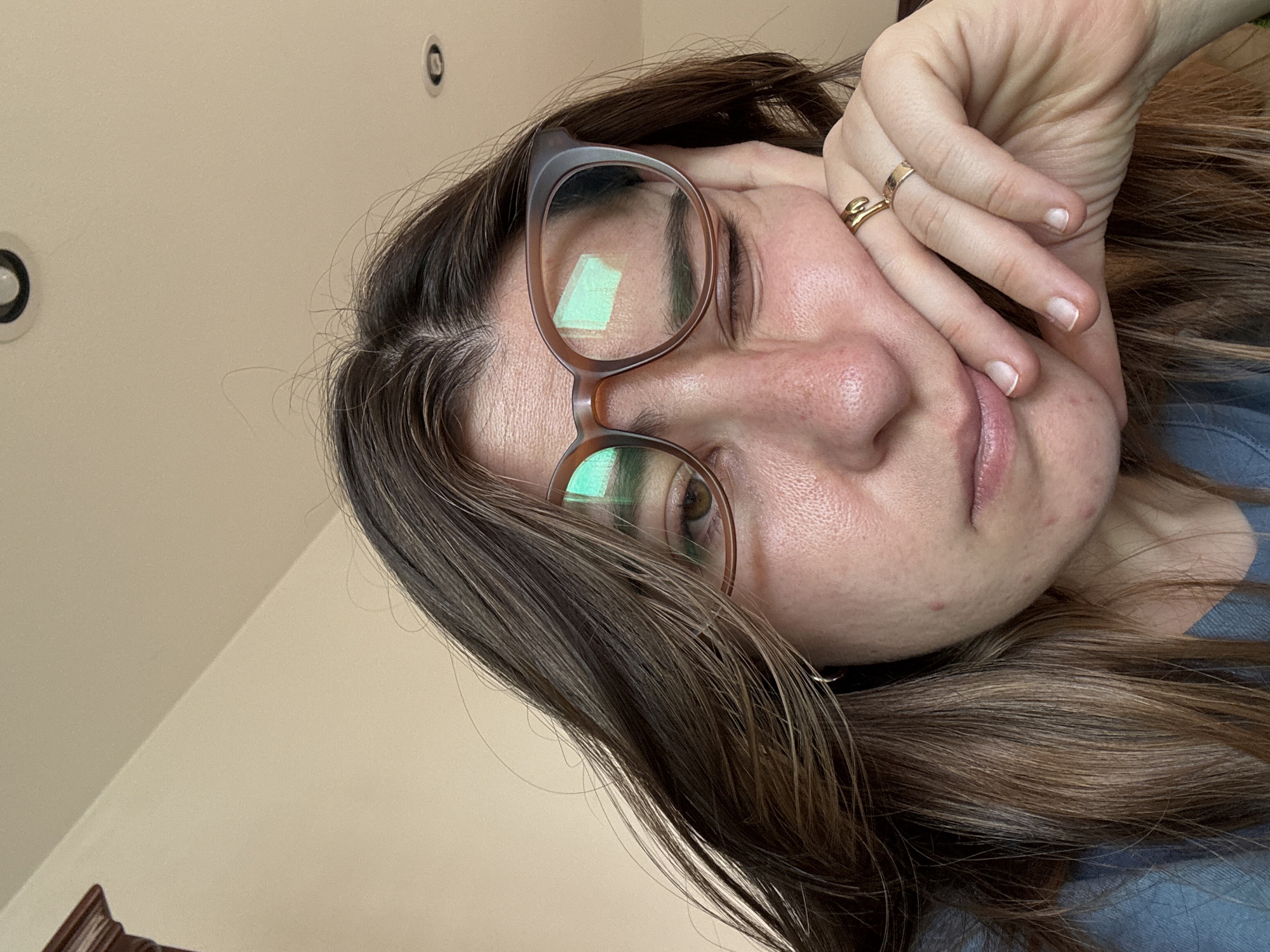 A close-up, sideways portrait captures a woman with dark brown hair and brown-framed glasses. Her right hand rests against her face, partially obscuring her forehead and cheek, revealing a gold-toned ring on her ring finger. Her expression appears pensive or slightly tired as she looks towards the camera. Rectangular reflections, possibly from a screen or light fixture, are visible in her glasses. Her skin shows natural texture and some minor blemishes, and her unpolished fingernails are visible. Her hair flows around her face and down the right side of the frame. The background consists of a light-colored, plain ceiling with two visible recessed circular lights. The image lacks any discernible text. This indoor scene is located in Puebla, Mexico.