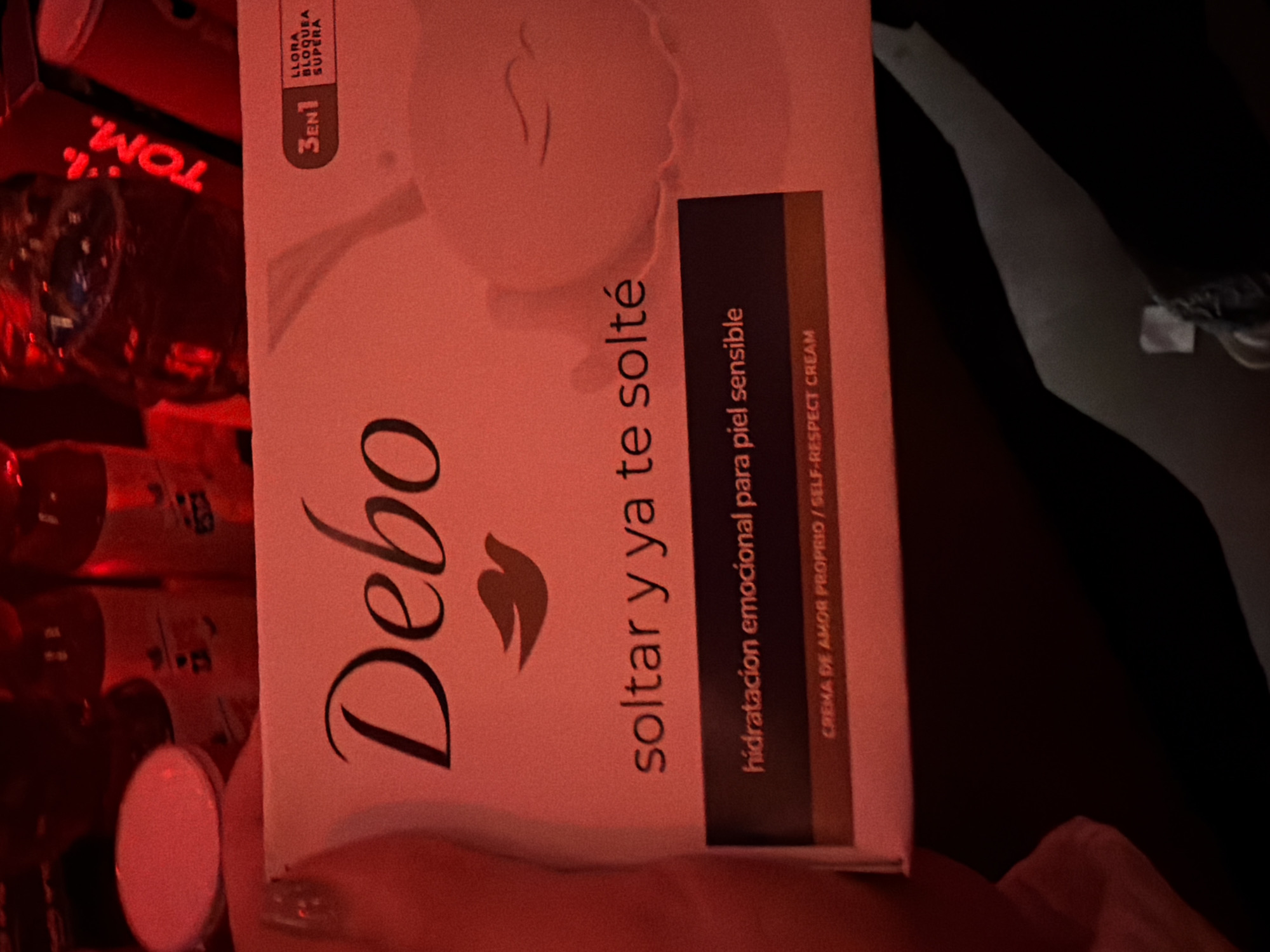 Here's a forensic description of the image:
The image shows a product box, partially obscuring the view. The box is white with pinkish undertones, bearing the brand name "Debo" in a stylized font. Below the brand name, text in Spanish reads "soltar y ya te solté," followed by "hidratacion emocional para piel sensible," and "CREMA DE AMOR PROPRIO / SELF-RESPECT CREAM". The box also has a "3EN1" label with the words "LLORA BLOQUEA SUPERA". The packaging is positioned in front of a cluttered background, featuring red-lit items, including a glass bottle bearing the name "TOM." A hand is visible, partially obscuring the bottom of the box.