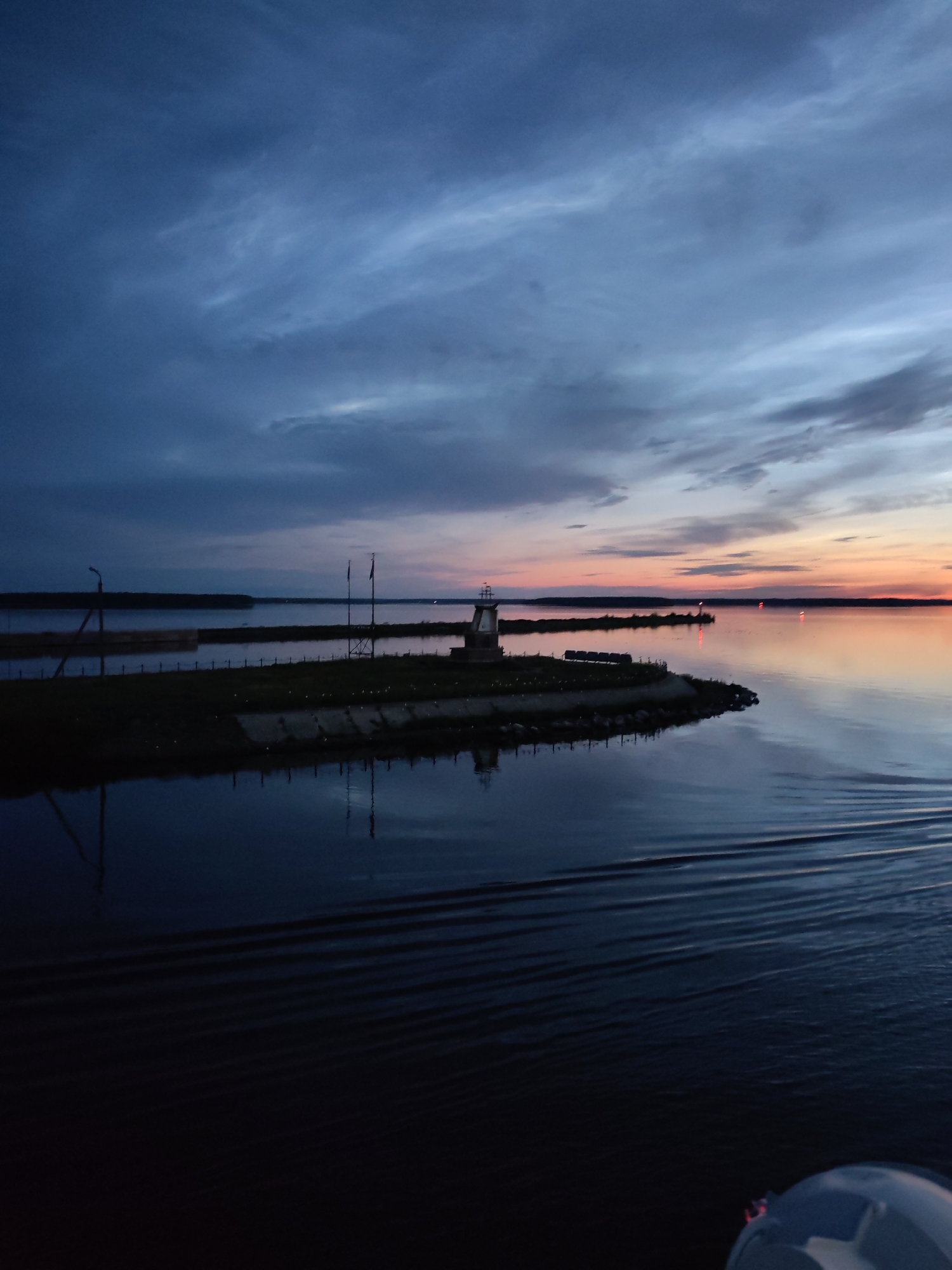 The image depicts a body of water, possibly a river or a lake, at dusk. The sky is overcast with dramatic clouds in shades of blue and grey, with a hint of orange and pink near the horizon, indicating the setting sun. In the foreground, the water is dark and reflects the sky and the surrounding landscape. There are visible ripples on the water's surface, suggesting movement, perhaps from a boat.
On the left side of the image, a dark silhouette of land with some vegetation is visible, with a tall, thin structure, possibly a lamppost, on the edge. In the mid-ground, a small, man-made peninsula juts out into the water. It is topped with a small structure that resembles a lighthouse or a memorial, and there are what appear to be flags or poles. The peninsula is lined with a low wall or embankment, and there are some scattered lights visible on the water in the distance.
The overall impression is one of a serene, yet dramatic, natural landscape during twilight. Given the information that the location is Sheksna, Russia, this could be a view of the Sheksna River or one of the nearby bodies of water. The scene captures the quiet beauty of the environment at the end of the day.