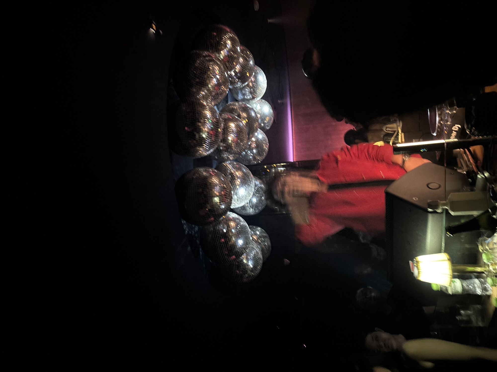 In the dim lighting of a nightclub in Thủ Đức, Vietnam, a DJ is visible behind a turntable, blurred by motion. Above, a cluster of disco balls hang from the ceiling, reflecting the ambient lights. In the foreground, a table holds bottles of drinks, a lamp with a white shade, and other miscellaneous objects. Several people are present in the scene, some in focus and others blurred by the low light and movement. The overall atmosphere suggests a lively night out.