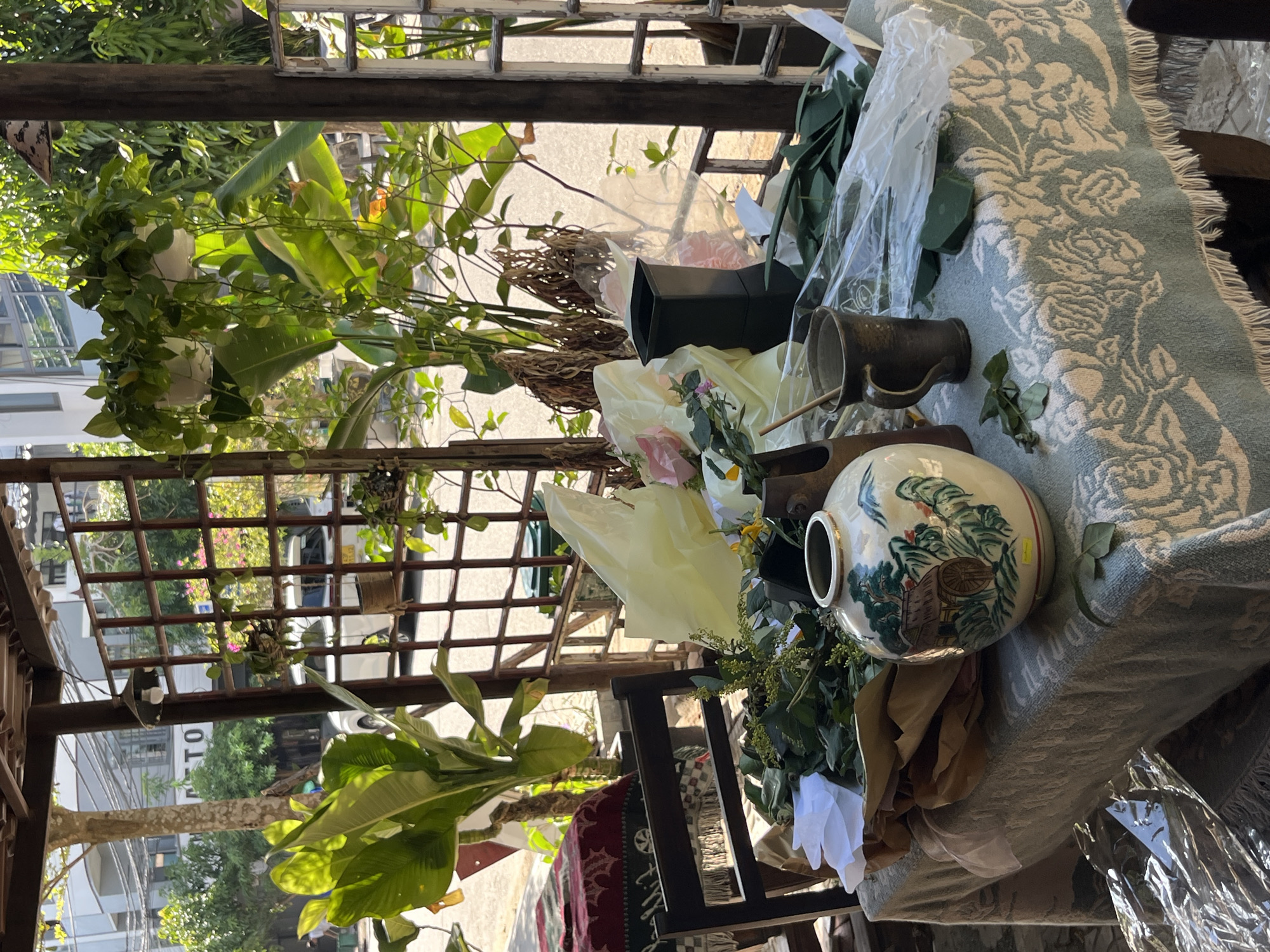 This image depicts a close-up view of a table covered with a light blue and cream floral patterned cloth, set outdoors under a covered structure. The table is laden with floral arrangement supplies, including a large, ornate ceramic vase painted with a traditional scene of a hut and greenery, various bunches of green foliage, a dark green floral foam block, a dark ceramic mug with a wooden stirring stick, and several sheets of light yellow and clear plastic wrapping. Behind the table, a wooden chair is partially visible. The background reveals a street scene in Đà Nẵng, Vietnam, with buildings, trees, a white car, and electrical wires under a bright, sunny sky. The overall impression is that of a workspace for flower arranging set up in a semi-outdoor, possibly residential or small business, setting in the city.