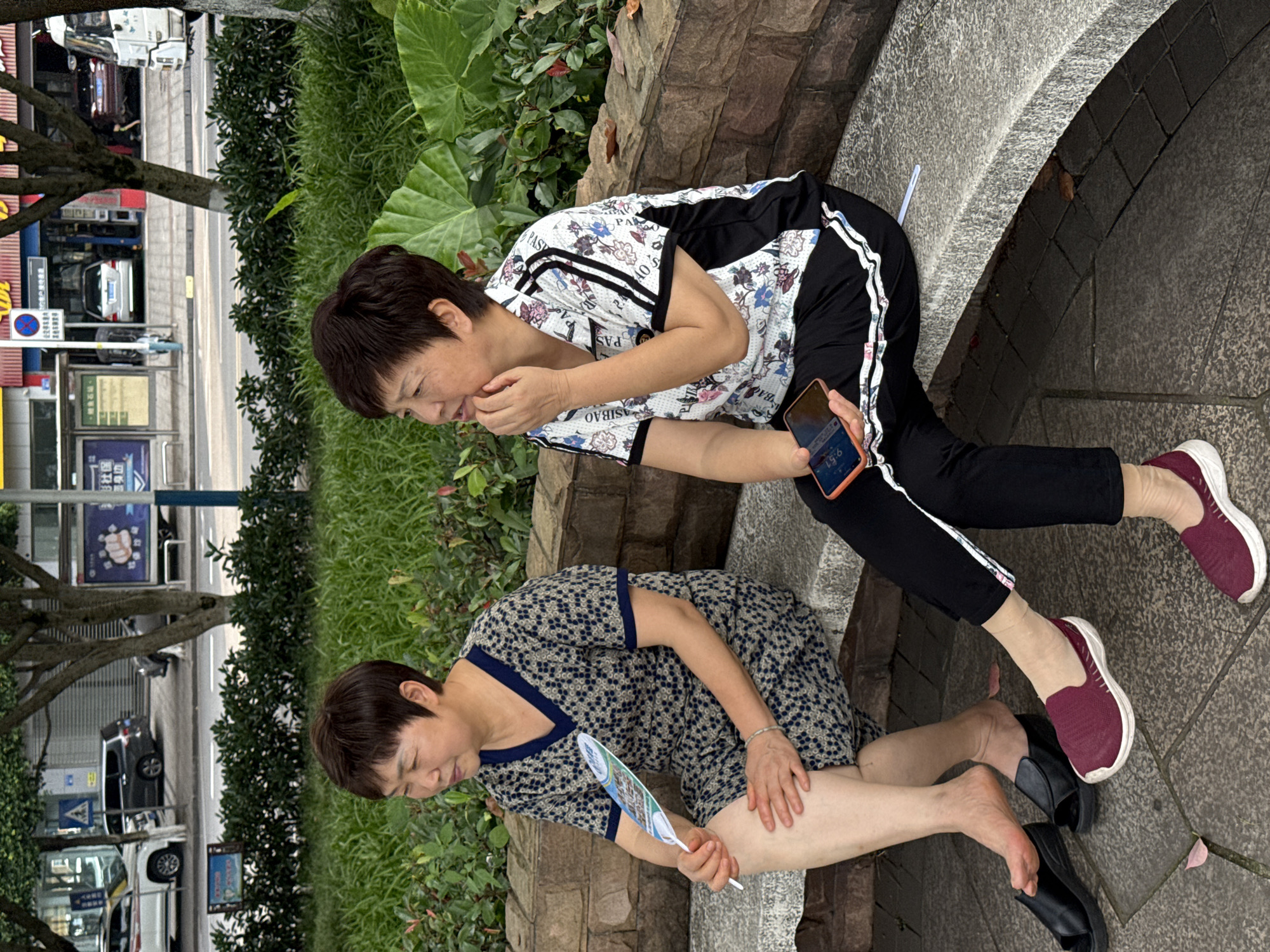 Two middle-aged Asian women are seated on a curved stone bench in a public green space in Jiangjin, China, with an urban street and buildings in the background.
The woman on the left wears a short-sleeved, knee-length dress with a dark blue V-neck and a patterned light-colored fabric featuring small dark blue circles. She has short, dark brown hair and a silver bracelet on her left wrist. She holds a white, feather-shaped fan with blue and green graphics in her right hand, while her left hand rests on her left knee. Her bare feet are visible, and she wears black open-toe sandals. She looks towards her left.
The woman on the right wears a short-sleeved collared shirt with a white base, black sleeves with two white stripes, and a multi-colored floral/abstract pattern. She also has short, dark brown hair. She wears black pants with a white stripe down each leg, light-colored socks, and burgundy athletic shoes with white soles. Her right hand is near her chin, and she holds an orange-cased smartphone displaying "9:51" in her left hand. She looks towards her left, in the same general direction as the other woman.
Behind the women is a stone retaining wall and dense green foliage, including grass, shrubs, and large-leafed plants. Beyond the green space is a paved road with a yellow solid line, indicating traffic flow. Several vehicles are visible on the road: a white SUV/car, a dark grey minivan/SUV, a silver sedan parked in front of a building, a red car, and a white bus/van. Commercial buildings line the street, one with a prominent red facade. Various street signs are present, including a pedestrian crossing sign and a no-parking sign, along with billboards displaying Chinese characters. Trees with green canopies are interspersed between the park area and the buildings. The lighting is overcast.