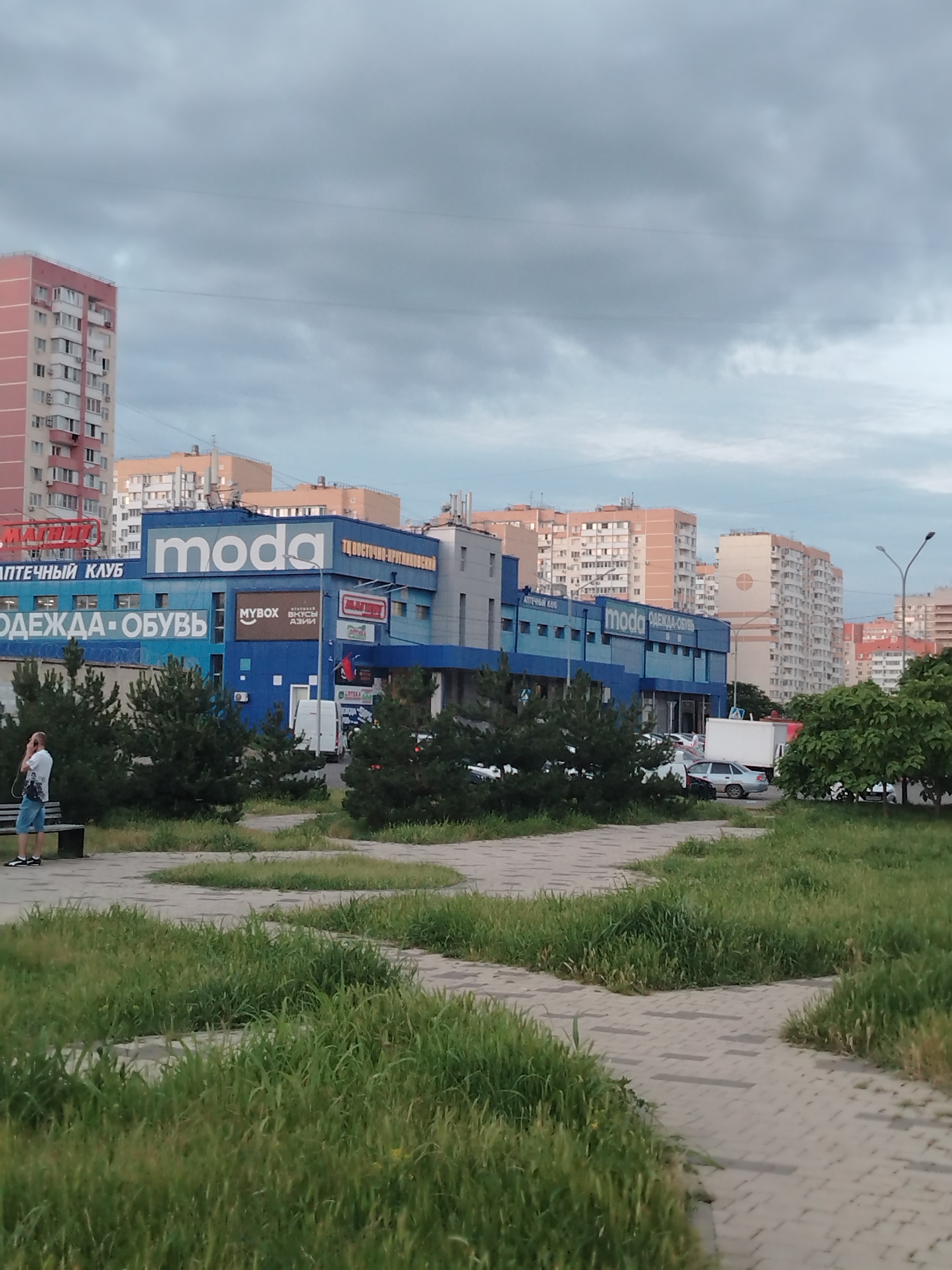 The image depicts a street scene in Krasnodar, Russia, on an overcast day. In the foreground, a paved walkway with patches of tall grass is visible. A person stands near a bench on the left side of the frame, looking at their phone. Behind them is a row of evergreen trees. Dominating the middle ground is a long, blue commercial building with various signs. Notable signs include "moda" in large white letters, "ОДЕЖДА-ОБУВЬ" (Clothing-Shoes), and "АПТЕЧНЫЙ КЛУБ" (Pharmacy Club). Several white vans and cars are parked in front of the building, suggesting commercial activity. In the background, several multi-story residential buildings with light-colored facades are visible, characteristic of urban architecture. The sky is filled with dense, gray clouds, indicating a possibility of rain. The overall impression is of a typical urban environment on a cloudy day.