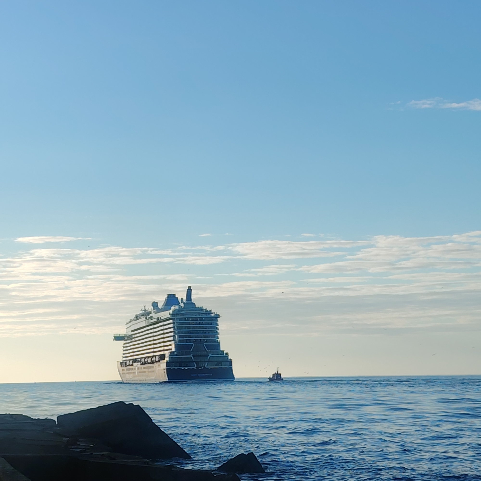 The image depicts a large cruise ship sailing on the Baltic Sea near Riga, Latvia. The ship, a multi-decked vessel with visible rows of cabins, is positioned towards the center-left of the frame and is accompanied by a smaller boat. The scene includes a body of water, presumably the sea, with visible waves and a rocky breakwater in the foreground. The sky is blue with scattered clouds, with indications of a bright, possibly sunny day. There is also an indiscernible amount of birds in the image.