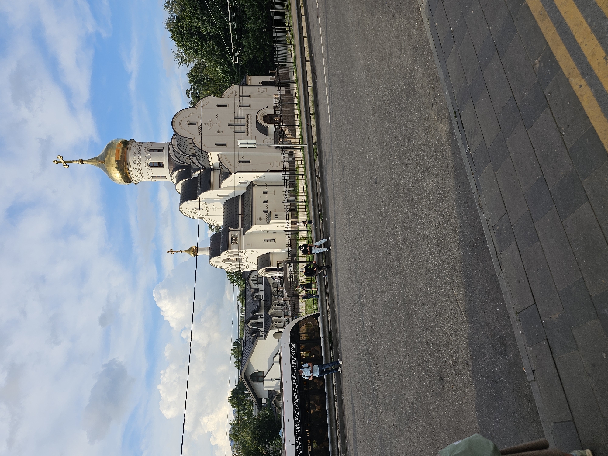 The image shows a modern white Orthodox church with a golden dome, located in Khimki, Russia. In the foreground, there is a paved road and a sidewalk with yellow markings. To the left, a modern tram is visible, with passengers inside and one person standing on the sidewalk next to it. Several other people are standing or sitting along a fence separating the sidewalk from the church grounds. Trees and a contemporary building with a dark roof are visible behind the church. The sky is partly cloudy.