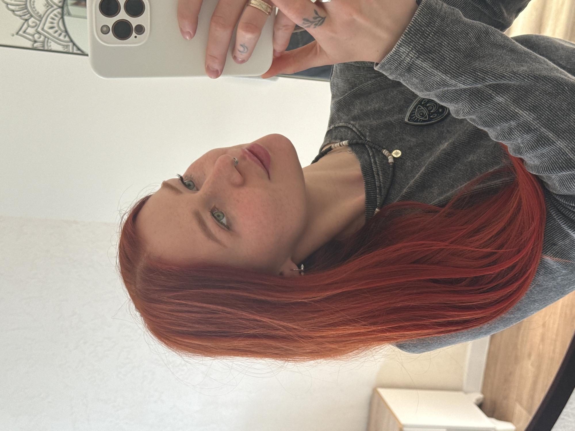 A selfie portrait features a young woman with fiery red hair and fair skin, her head tilted slightly to the left. She has freckles scattered across her face and piercing green eyes. A silver hoop is visible in her left nostril. She's wearing a gray corduroy jacket with a decorative silver pin and a beaded necklace. She's holding a white smartphone with multiple camera lenses. Her fingers are adorned with rings, including a delicate tattoo of a leaf branch on her finger. The background is a slightly blurred interior, possibly a hallway or bedroom, with white walls and glimpses of wooden furniture. There is nothing to suggest that the image was taken in the city of Rostov-on-Don, Russia.