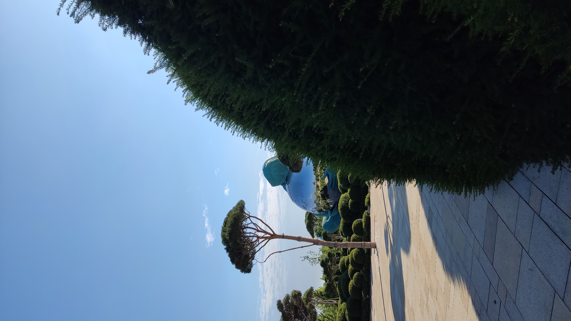 The image depicts a large, shiny, spherical sculpture partially obscured by lush green bushes and a tall evergreen tree in the foreground. The sculpture appears to be a stylized representation of a person holding a globe, made of a highly reflective material that mirrors the surrounding environment and the blue sky. The setting is a park or garden, with neatly trimmed shrubs and paved pathways. In the background, more trees and a clear blue sky with scattered clouds are visible. This scene is located at the "Galitsky Park" in Krasnodar, Russia.