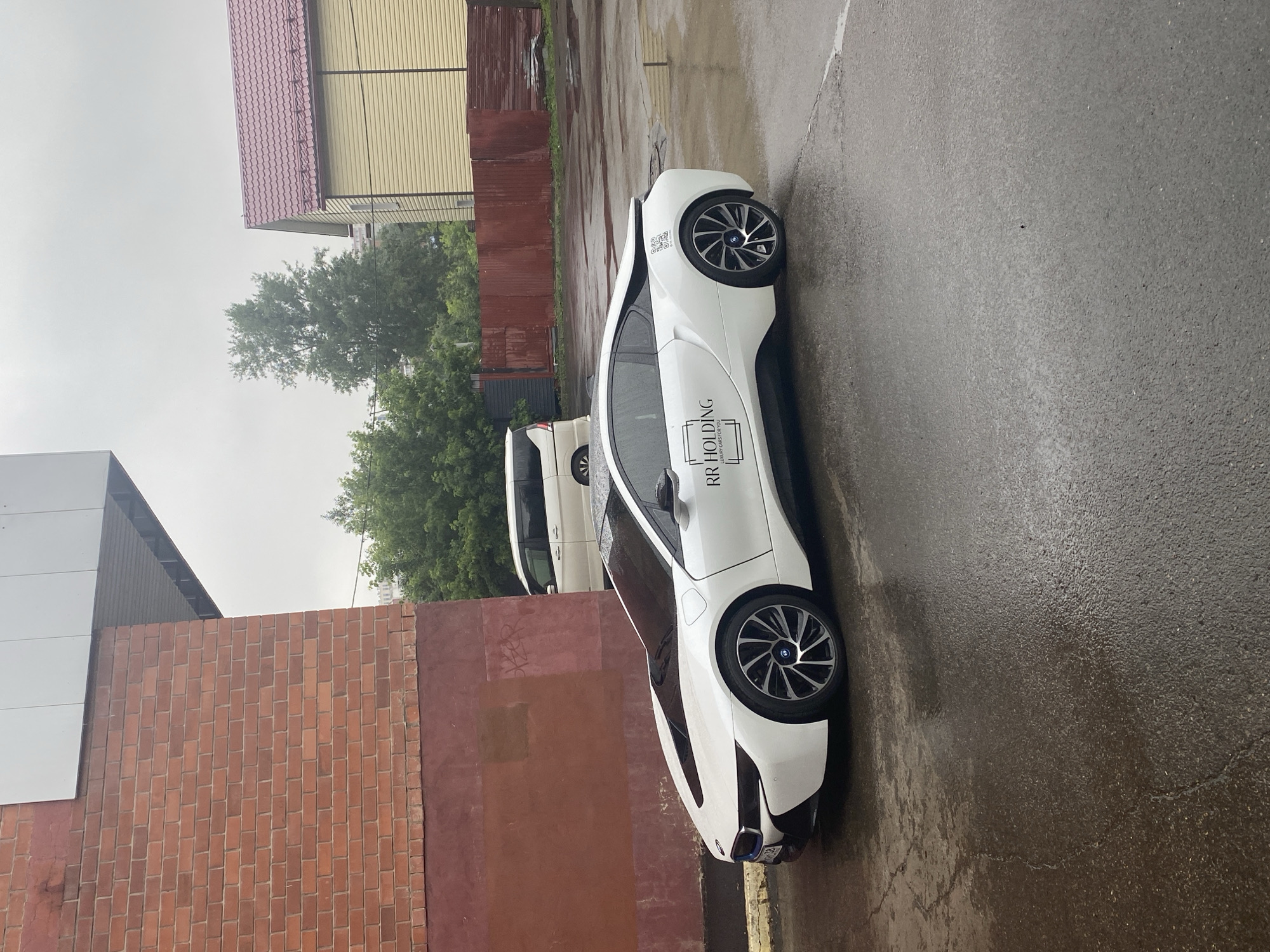 An exterior, eye-level, full shot depicts a white BMW i8 parked on a wet asphalt surface. The car is positioned to the right of a red brick building on the left side of the frame. The building features a dark red base and a lighter red brick facade above. A white awning extends from the building over the car. In the background, behind a red corrugated metal fence, are trees and two buildings: one with a yellow exterior and a red roof, and a white van. The sky is overcast and grey, suggesting a rainy or damp day. The wet asphalt reflects the surrounding environment. A logo with the text "RR HOLDING" and "LUXURY CARS FOR YOU" is visible on the side of the car. A QR code is also present on the rear quarter panel of the car. The scene appears to be an outdoor parking area, possibly in a commercial or industrial zone. The overall mood is subdued due to the weather and lighting conditions. The image was taken in the city of Irkutsk, Russia, based on context provided elsewhere.