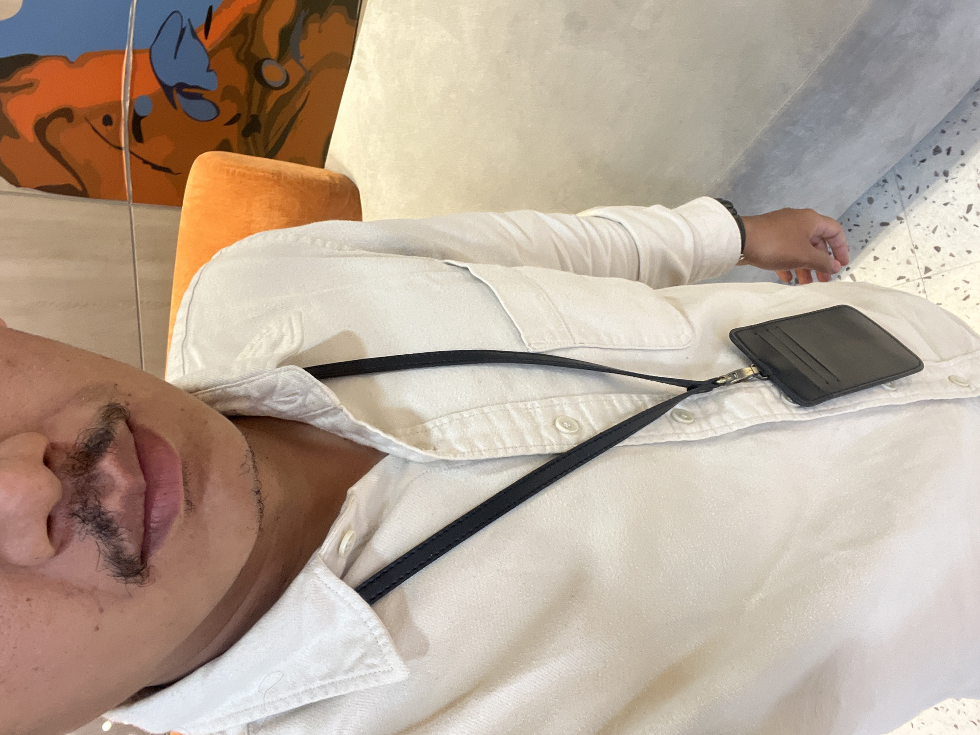 A close-up, medium shot shows a person from the chest up, wearing a light-colored collared shirt. A black lanyard with a black cardholder is visible around their neck. The person has a mustache and a goatee. The background includes an orange upholstered chair and a portion of a colorful abstract painting. The floor has a terrazzo-like pattern. The lighting appears to be indoor ambient light. There are no clear indications of the specific location within West Jakarta, Indonesia.