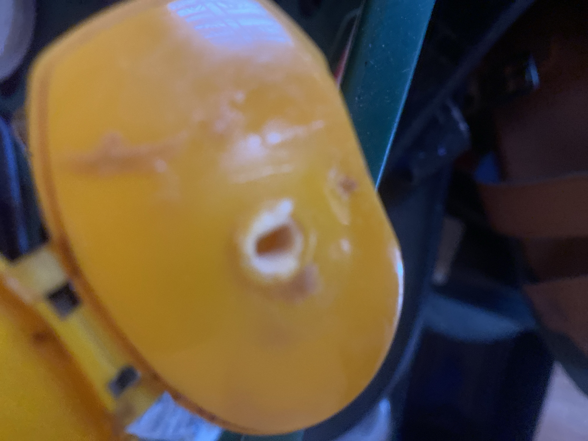 The image shows a close-up of a bright yellow object, possibly a plastic toy or part of a larger item, with a hole in the center. The surface of the yellow object appears to be somewhat worn, with smudges and a few light brown marks. The hole in the center is irregular in shape and has a lighter, possibly whitish, rim. The object is set against a dark background with hints of other objects and colors, including a dark green element on the right and a darker brown material. There are also some blurry elements of blue and yellow in the lower left. The lighting creates a slight glare on the yellow surface. No people are visible, and the scene appears to be an indoor setting, possibly a cluttered area or a workshop, given the presence of various materials and the close-up focus on a specific object. There is no discernible text in the image. The context provided of "Jalingo, Nigeria" does not offer any specific visual cues within the image itself to confirm or deny this location.
