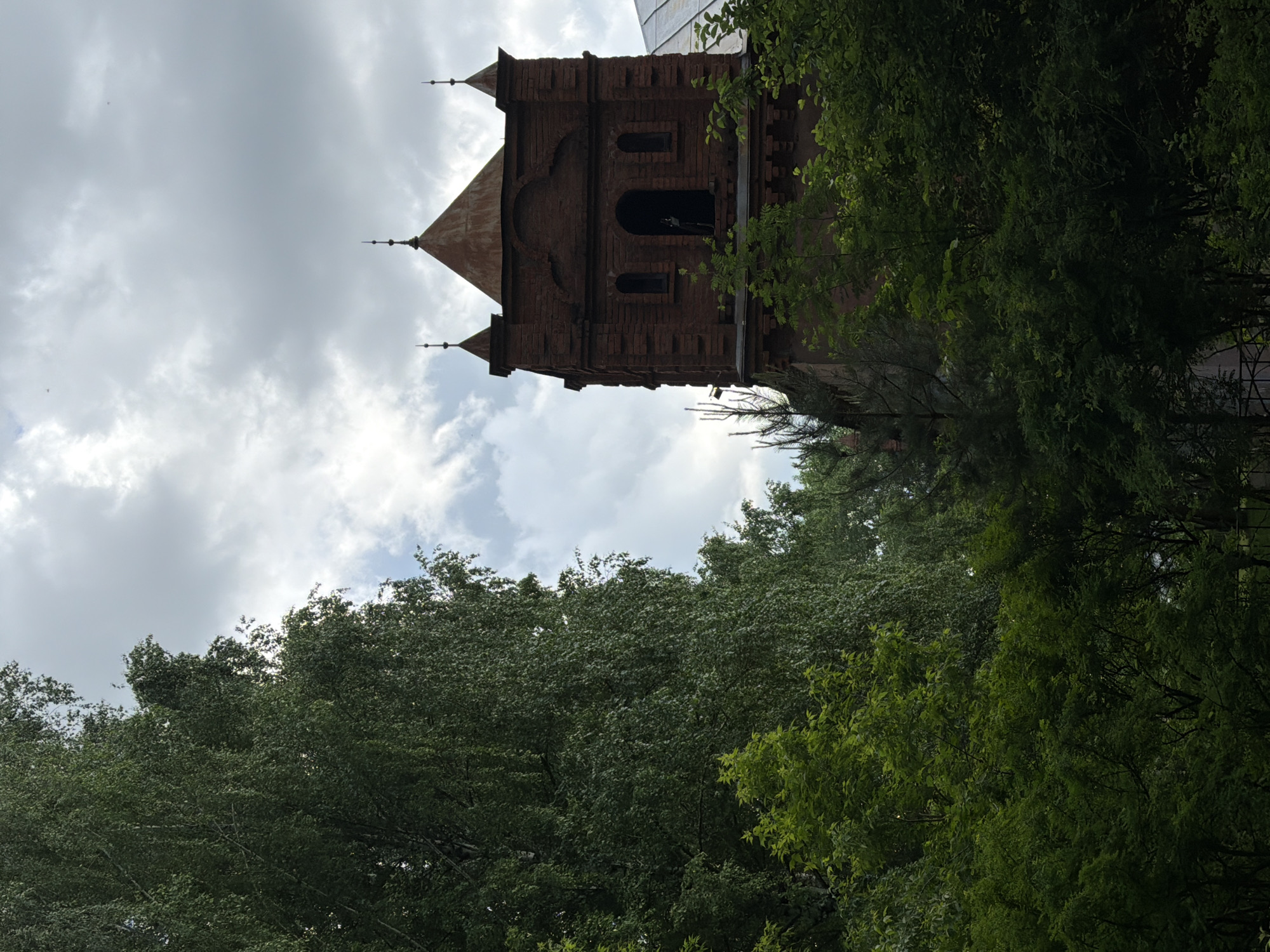 The image depicts the Votkinsk Technical College building in Votkinsk, Russia. The building, constructed of red brick, features a tower with a pointed roof and decorative turrets. A section of green trees borders the bottom of the image. The sky behind the building is cloudy.
