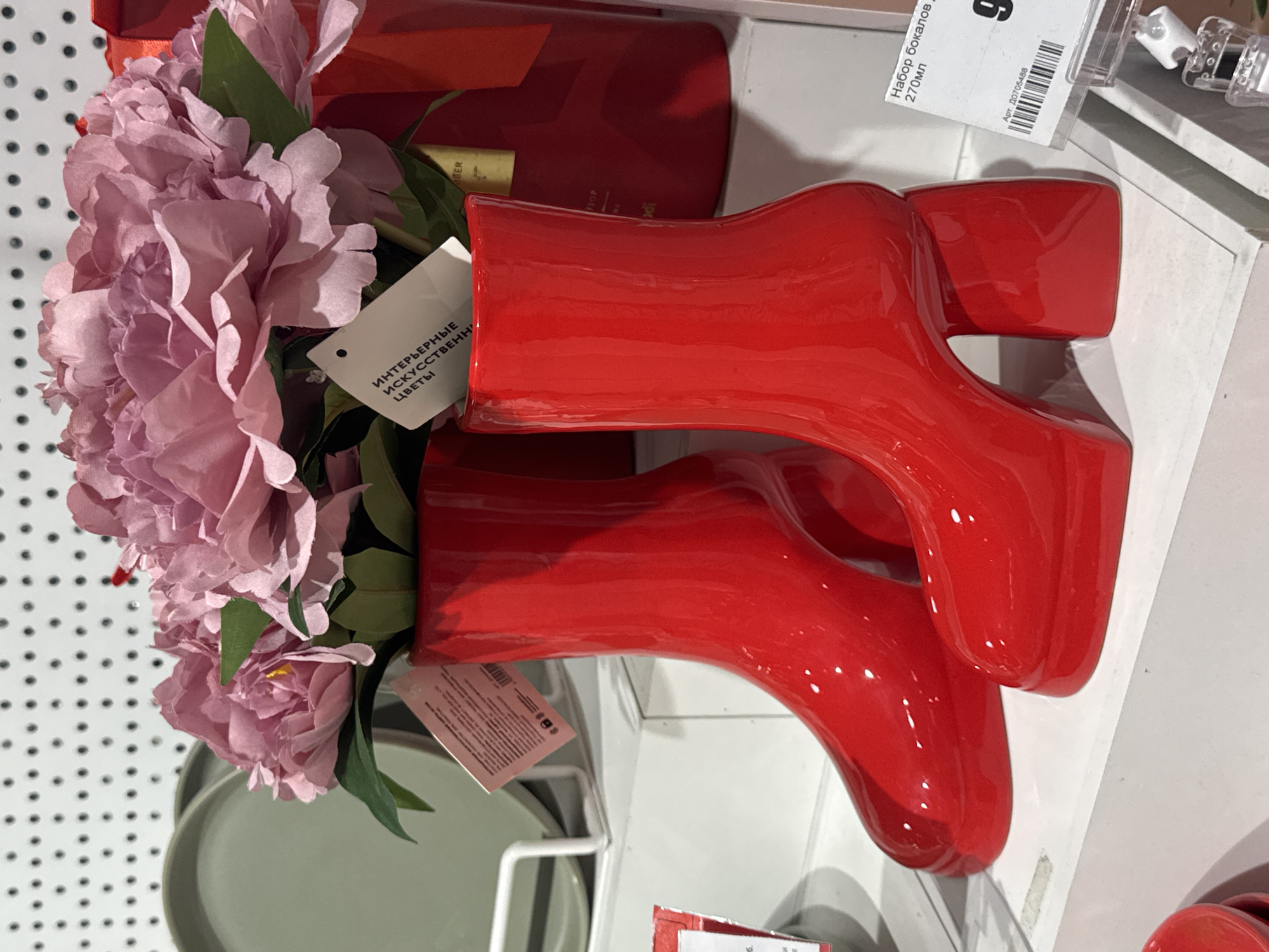 The image displays a close-up view of two glossy red vases shaped like high-heeled boots, filled with artificial pink peonies. The vases are positioned on a white shelf against a white pegboard background. A retail price tag, featuring Cyrillic text and a barcode, is visible on the right side, indicating the product is "Набор бокалов, 270мл" (Set of glasses, 270ml) and has an item number "Арт. Д0705488". The setting appears to be a retail environment, possibly a home decor store or a department store. There are no people visible in the frame, and the lighting suggests an indoor setting with artificial illumination. The context of Ufa, Russia, is not directly evident from the visual information within the image itself, but the Cyrillic text on the tag points to a Russian-speaking region.
