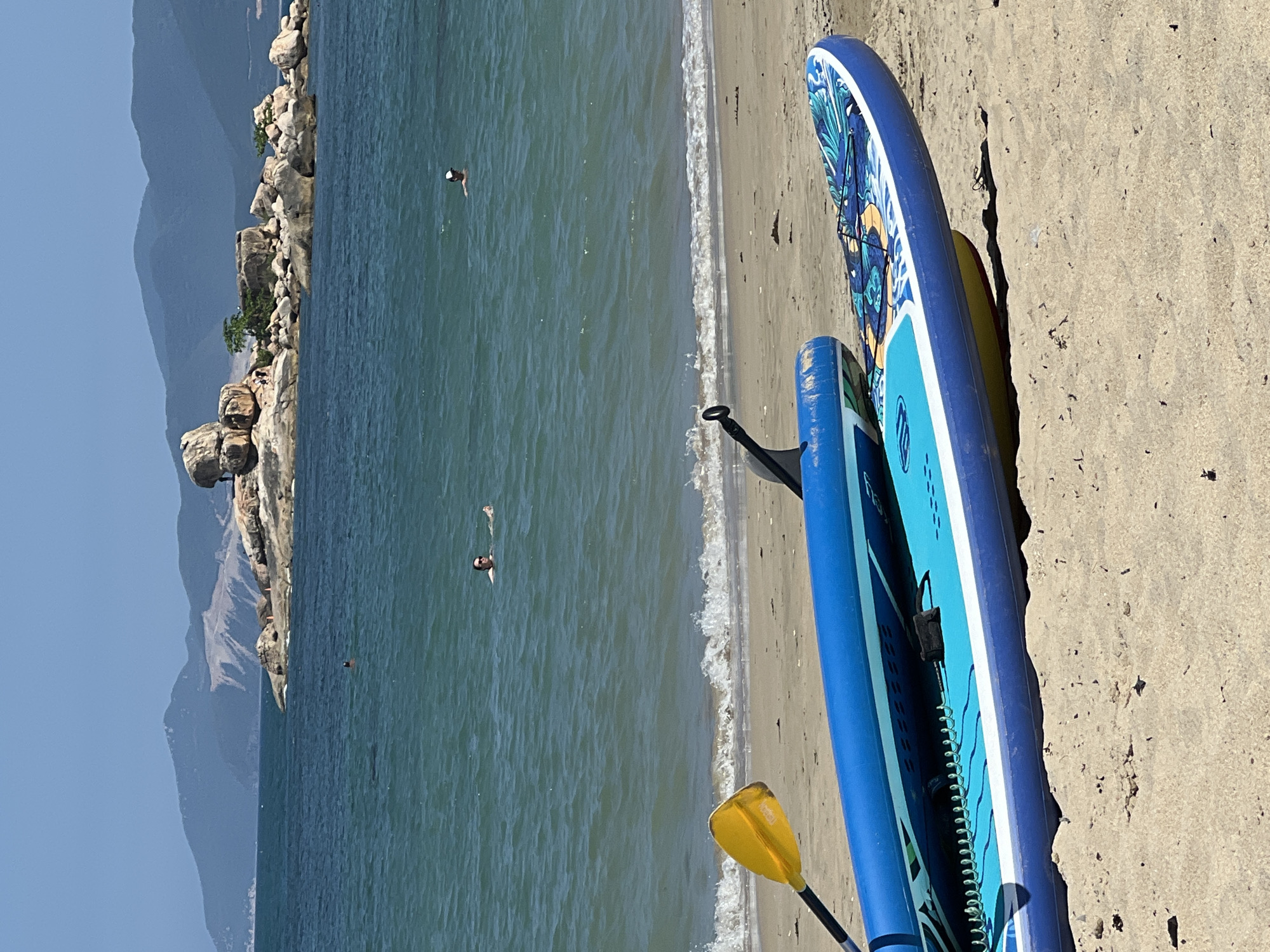 The image captures a coastal scene likely in or near Bắc Nha Trang, Vietnam, based on the distinctive rocky headland and mountain backdrop. In the foreground, two paddleboards rest on a sandy beach. One board is blue with white and decorative accents, and partially obscures a second, smaller blue board. A yellow paddle rests near the boards. The clear, turquoise water occupies the middle ground. Two individuals are seen swimming in the water. The background features a rocky headland with some vegetation and, in the distance, a mountain range is visible under a clear, blue sky. The overall lighting suggests a bright, sunny day.