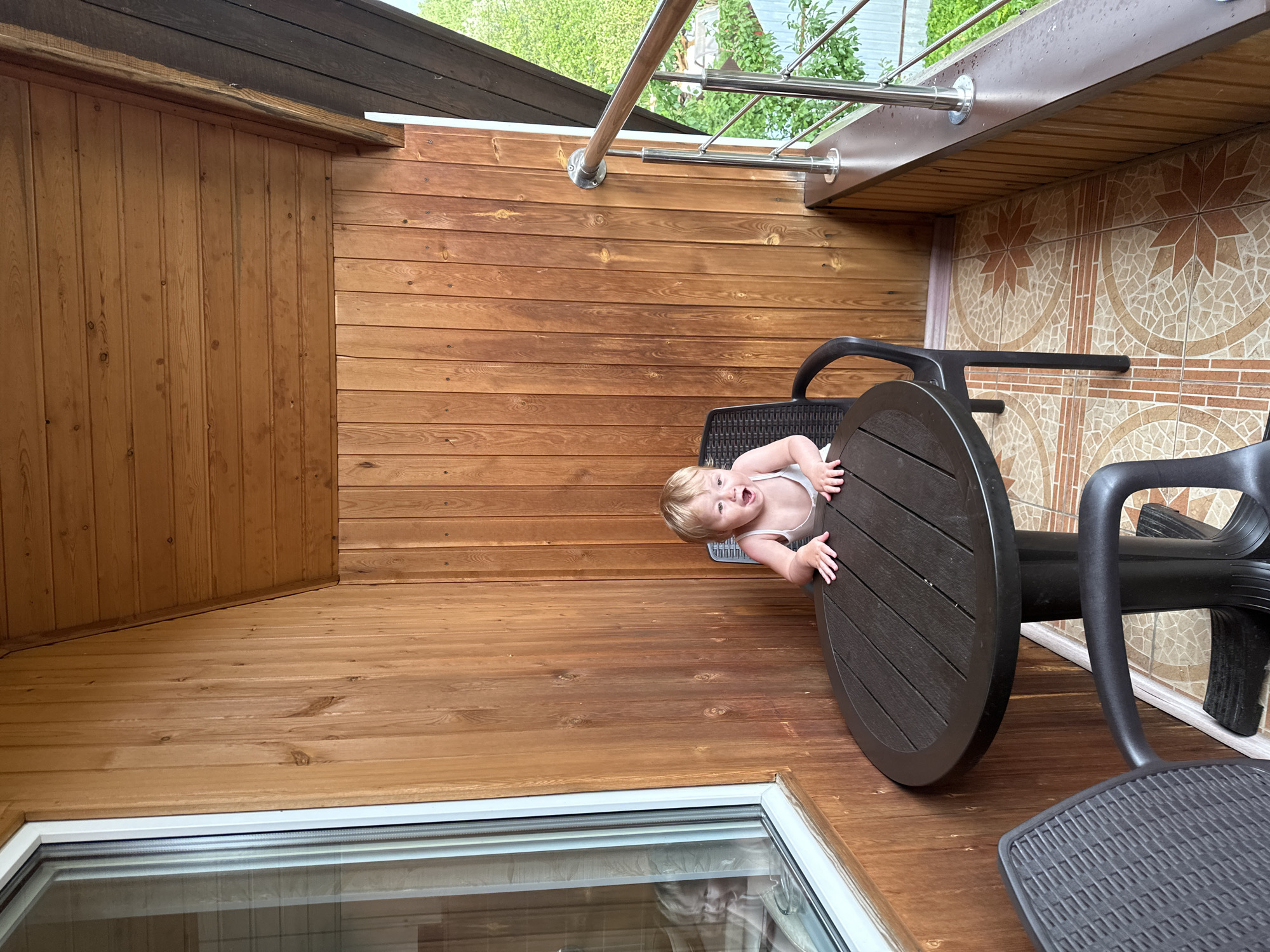 A young child with blonde hair and an open mouth is seated at a round, dark-colored table on a wooden balcony in Nizhny Novgorod, Russia. The child is wearing a white tank top and has their hands on the table. To the left of the child, a large window reflects another image of the child. The balcony has wooden walls and a tiled floor with a starburst pattern. A metal railing with a wooden top runs along the edge of the balcony, overlooking a green outdoor area.