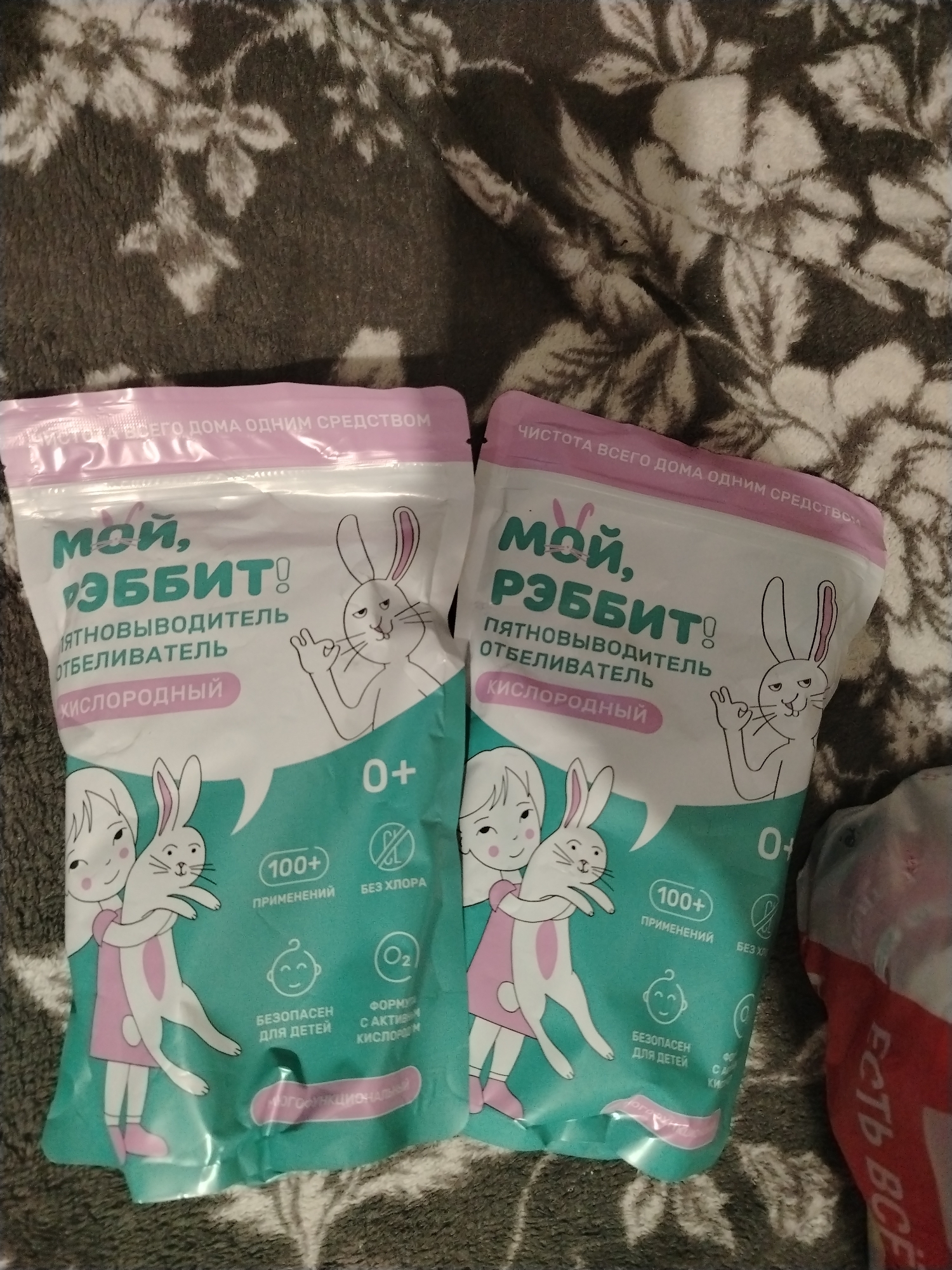 The image shows two pouches of laundry stain remover on a patterned surface. Each pouch is white with a pink top and features cartoon illustrations, including a rabbit. The text on the pouch is in Russian and includes the brand name "Мой, Рэббит!" (My Rabbit!), followed by the product description "Пятновыводитель отбеливатель кислородный" (Oxygen stain remover bleach). The pouches also display symbols indicating that the product is safe for children and suitable for multiple uses. A small portion of a red-and-white package is visible on the right side of the image.
