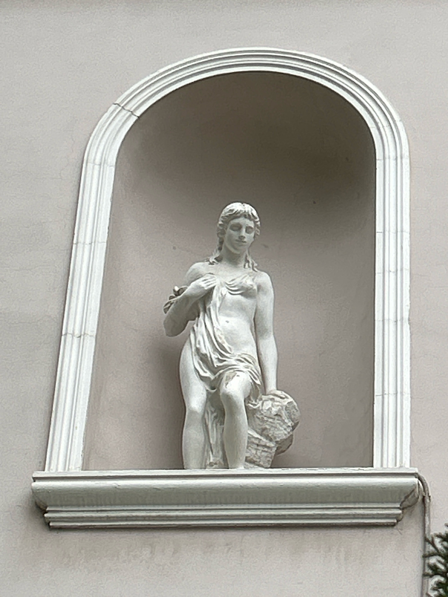 The image shows a white statue of a female figure in a niche on a building in Moscow, Russia. The statue depicts a woman with her arm over her chest, draped in fabric, and holding a bundle of what appears to be rock or vegetation at her side. She is positioned within an arched niche that has decorative molding. The niche is set into a light-colored wall. The statue and niche are illuminated by natural light, casting subtle shadows. The overall aesthetic suggests classical or neoclassical architectural influence.