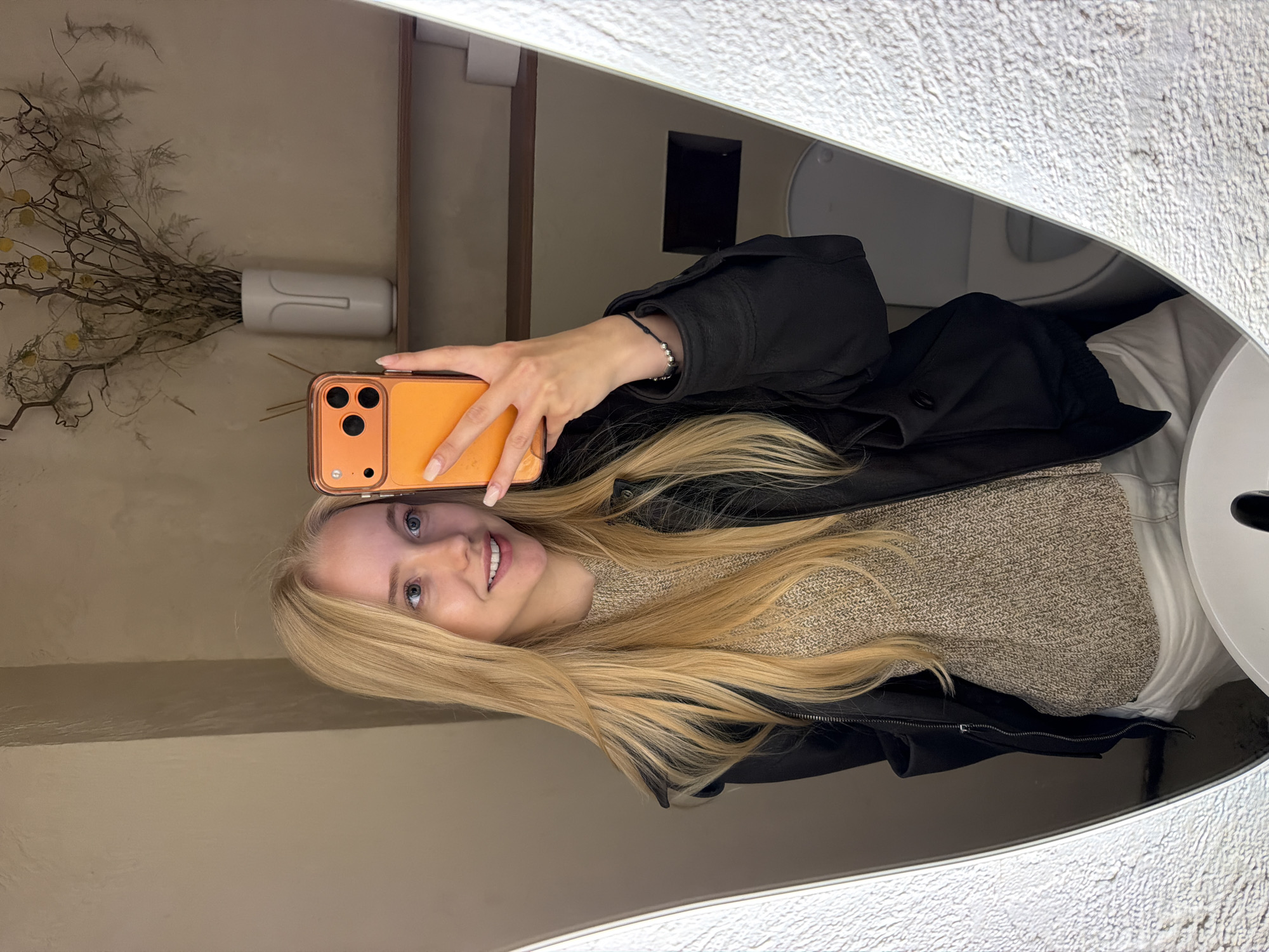 A young blonde female, appearing to be in her late teens or early twenties, takes a mirror selfie in a bathroom located in the city of Unknown, Unknown. She has long, straight, light blonde hair, fair skin, and blue eyes, and is smiling with her mouth slightly open. She wears a black zip-up jacket over a light brown knitted sweater and white or light-colored pants. A dark, beaded bracelet is visible on her right wrist. Her right hand holds an orange smartphone with a clear protective case, revealing its triple camera array on the back. The mirror's frame has a prominent, irregularly textured, light grey border on both the left and right sides. Reflected in the mirror behind the subject is a light beige-colored wall, a wooden shelf, and a partial view of a white toilet bowl. On the shelf, a white minimalist vase containing dried branches and yellow spherical floral elements is present, alongside a small container with dark diffuser sticks to the left. To the right of the vase, two white rolls of toilet paper are stacked. The overall environment suggests a clean, modern bathroom with neutral tones.