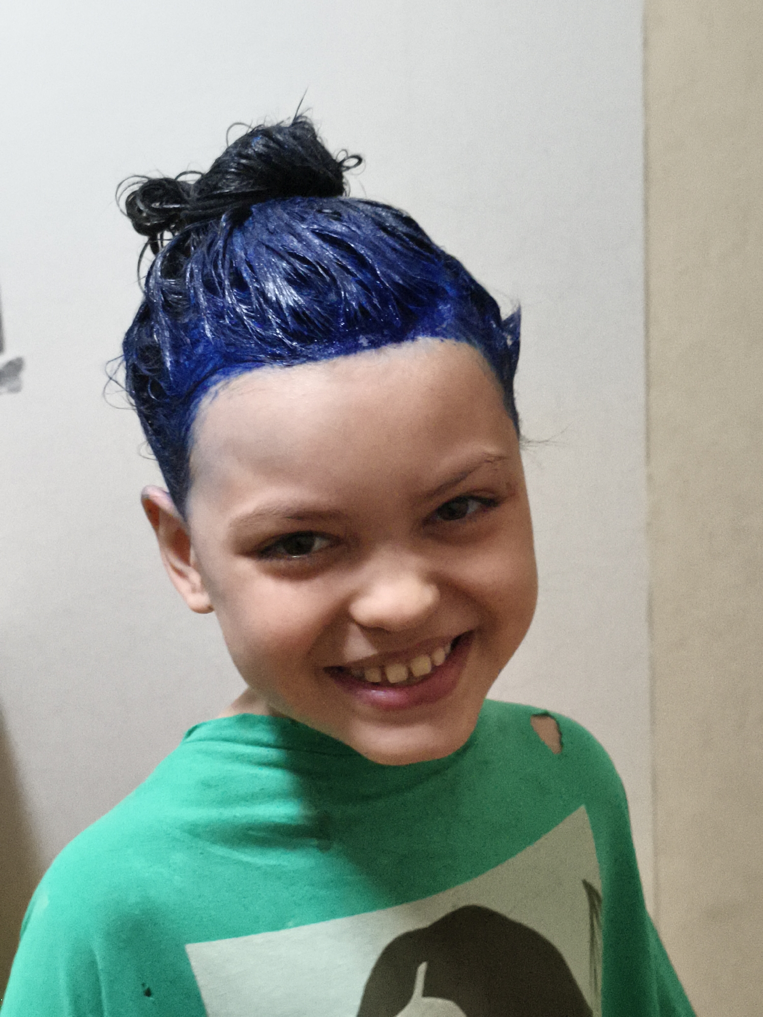 A young person with blue-dyed hair, styled in a messy bun, smiles at the camera. They are wearing a green t-shirt with a graphic print partially visible. The hair appears wet, suggesting it's in the process of being dyed or washed. The background is a plain, light-colored wall, indicating an indoor setting, likely a home. The lighting is even and appears to be from artificial sources. The expression on the person's face is joyful and playful. There is no visible text in the image. The context of Chaikovsky, Russia is provided but not visually evident in the image itself.