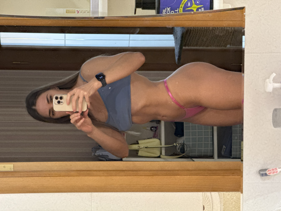 The image displays a mirror selfie of a young woman, rotated 90 degrees clockwise. The woman, with long, straight light brown hair, is looking directly at the camera through her smartphone. She is holding a light-colored iPhone with a triple-lens camera array in her right hand, capturing her reflection in what appears to be a large mirror.
She is wearing a slate-blue sports bra or crop top and bright pink, minimalist bikini-style bottoms or a thong. A black smartwatch is visible on her left wrist. The woman's physique appears athletic, with visible abdominal definition.
The setting is indoors, likely a bathroom or changing area, reflected in the mirror. Behind her, various objects are visible on shelves and ledges. On the top shelf, a white rectangular container with text "PLAY XL" can be seen, along with a purple bottle that shows partial text "CURL CK". Further back, there are glimpses of what looks like a grey storage unit or tiled wall, and a light-colored handbag. To the far right edge of the mirror, a white soap dispenser and a toothbrush holder with two toothbrushes are visible, further suggesting a bathroom setting. The frame of the mirror is made of wood. The overall context for the image is Saint Petersburg, Russia.