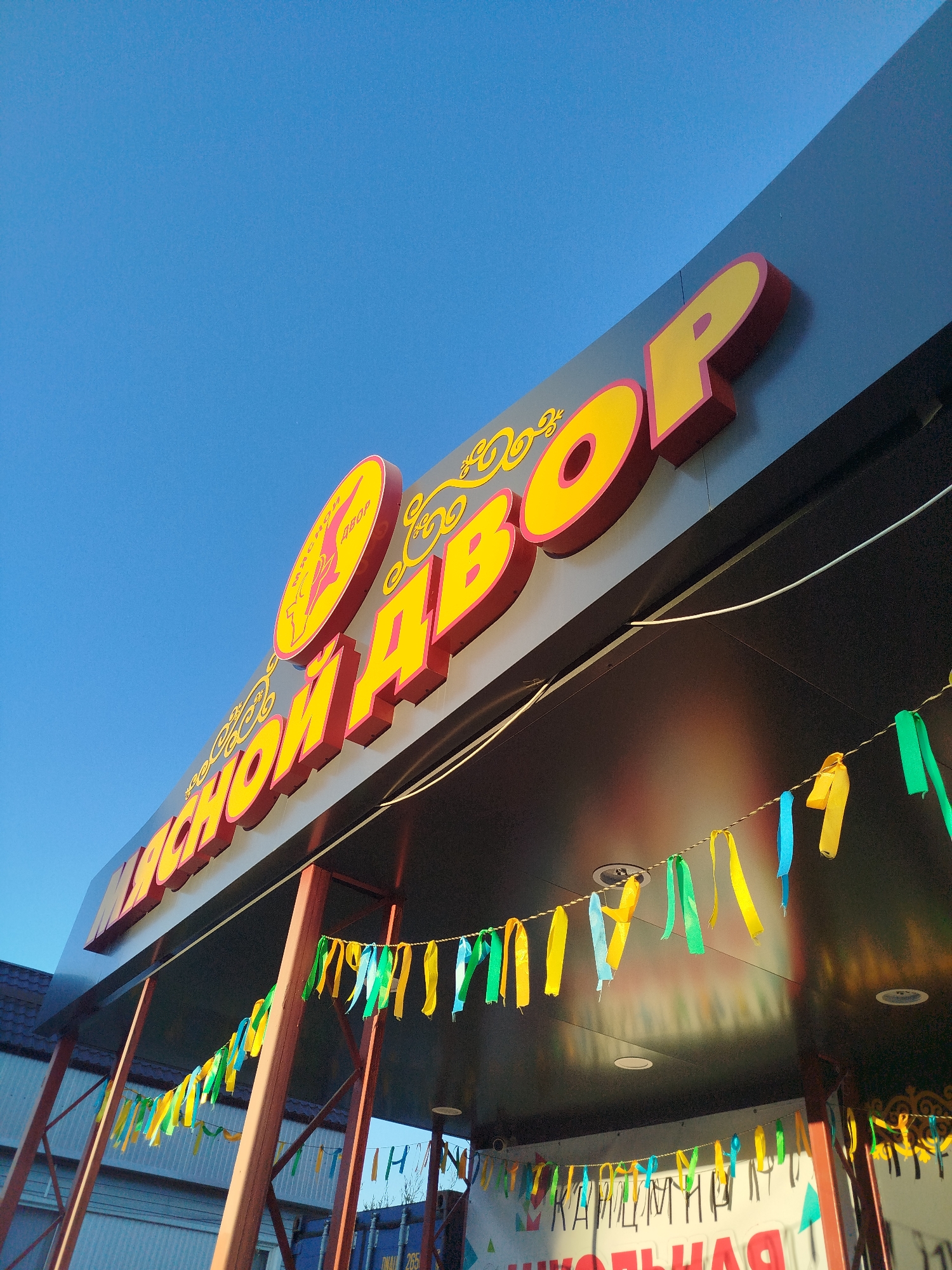 An outdoor low-angle shot captures a bright blue sky above a dark fascia with a prominent yellow and pink sign. The sign is three-dimensional with bold, rounded letters spelling out "КРАСНОЙ ДВОР" which appears to be a Russian phrase. To the left of the main sign, a circular emblem with a stylized image is visible. Below the fascia, a string of colorful ribbons in green, yellow, and blue hangs, festooned between decorative red-brown metal supports. A partial view of a white building and a blue shipping container is visible in the background. The scene is likely in Mirny, Russia, given the Cyrillic alphabet used in the signage. There are no people or animals visible in the image.
