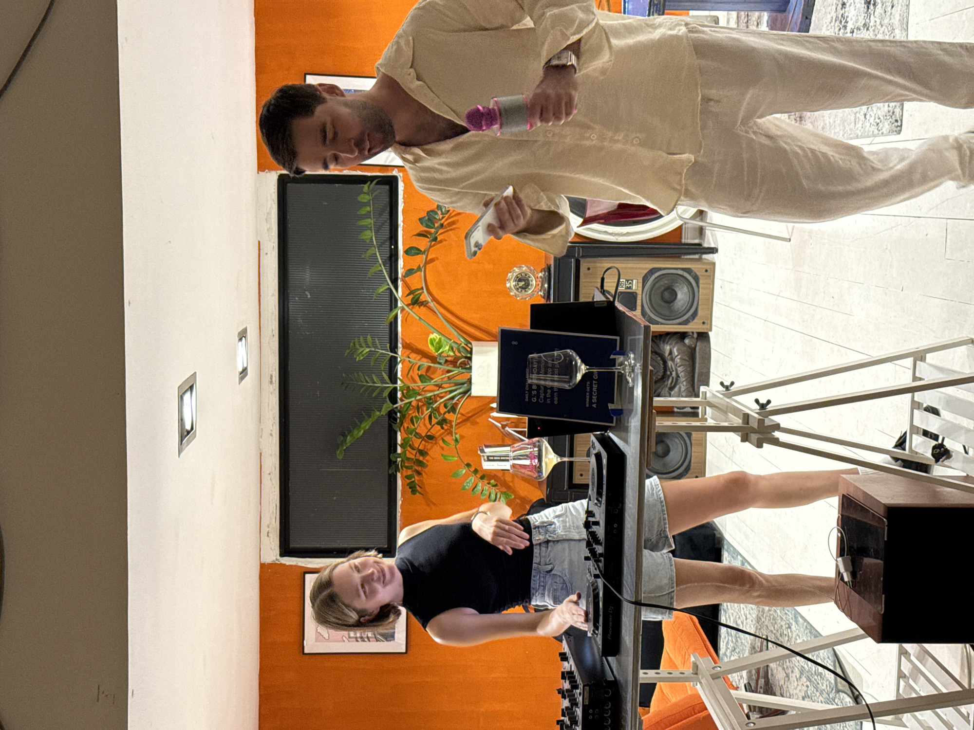 In Tel-Aviv, Israel, a man and a woman are in an indoor room with an orange wall. The woman, wearing a black sleeveless top and denim shorts, stands to the left of a DJ setup, with her hands on the turntables. She has short blonde hair and is smiling. To her right, a man in a light-colored open-collared shirt and matching pants holds a smartphone in his left hand and a pink karaoke microphone in his right. He is looking down at the phone. In the background, a plant sits in a pot on a table, and there are speakers and other objects. The scene appears to be a home or a casual gathering space.