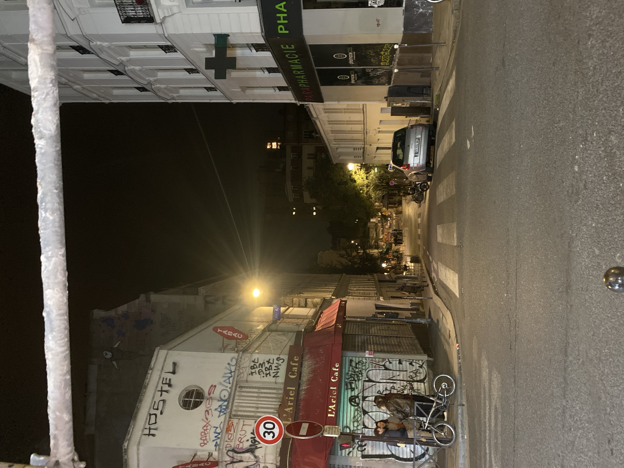 The image depicts a street scene in Paris, France at night. The view is looking down the street, with buildings on either side. On the left, a building with "HOSTEL" written on it and graffiti, and a cafe named "L'Ariel Cafe" with red awnings and signs. A speed limit sign showing 30 is visible. Bicycles are parked near the cafe. On the right, a building with "PARAPHARMACIE" written on it, which is a pharmacy. A car is parked on the street, and several people are walking in the distance. The street is illuminated by street lights. A white textured pole appears in the front.