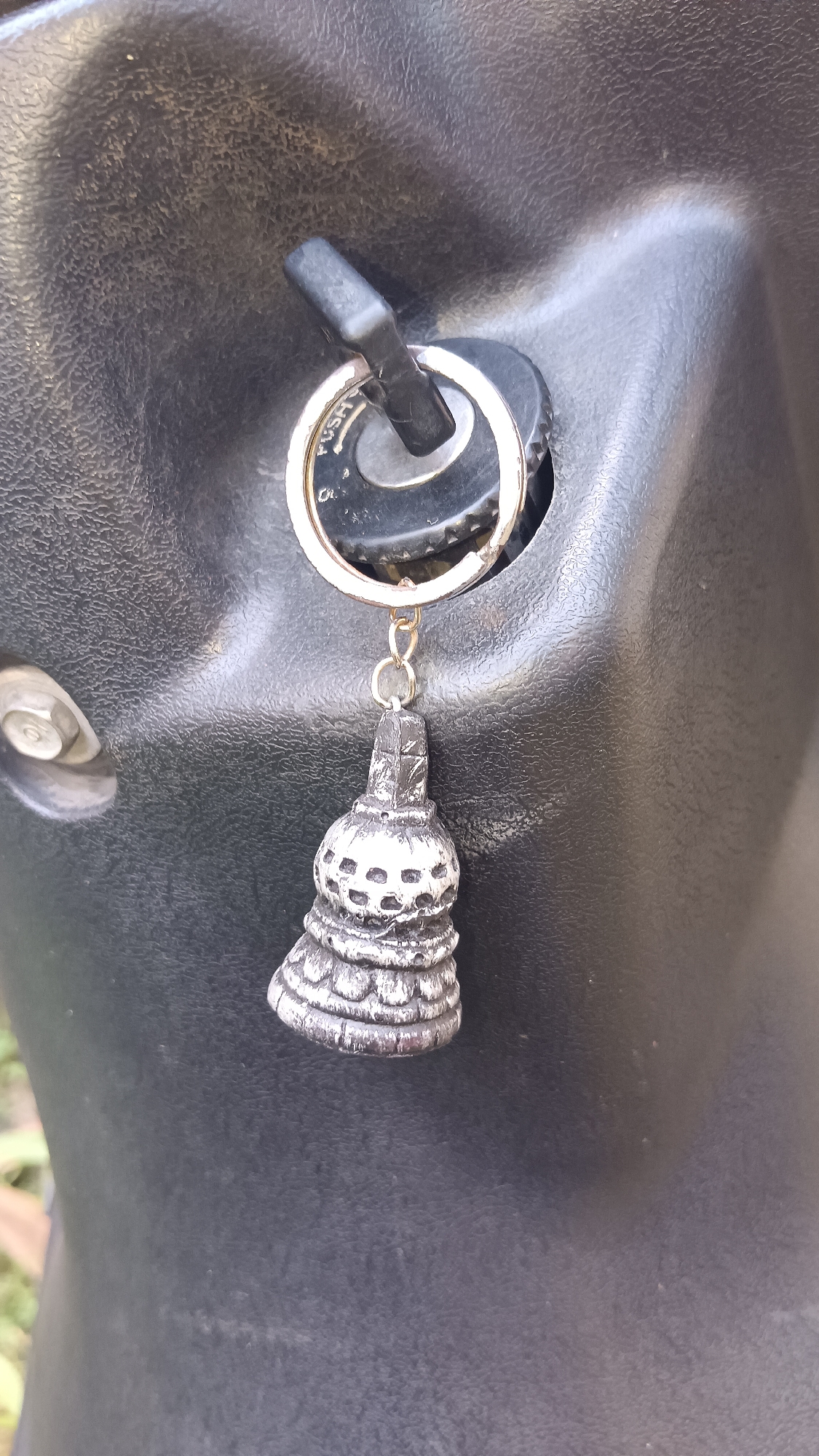 A close-up, eye-level shot focuses on a key inserted into a black ignition switch. The key has a circular metal keychain with a small, silver-colored charm dangling from it. The charm is intricately designed, resembling a miniature bell with multiple tiered layers and circular cutouts. The ignition switch itself is partially obscured by the key. The background is dominated by the textured black surface of what appears to be a vehicle, possibly a scooter or motorcycle. A portion of a bolt or screw is visible on the left side of the frame. The lower left corner shows a hint of greenery, suggesting an outdoor environment. No people or other distinguishing landmarks of Kemang, Indonesia are visible.