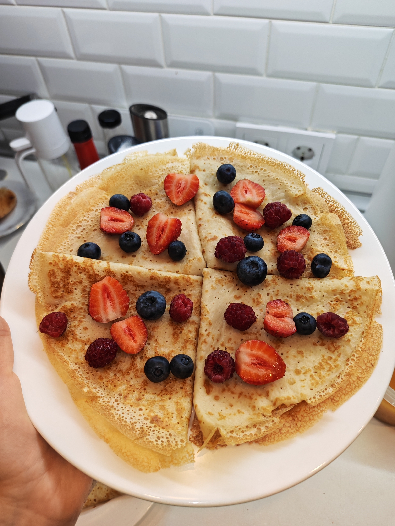 A close-up, overhead shot shows a white plate filled with four crepes, each cut in half, creating a circular arrangement on the plate. The crepes are golden brown and sprinkled with fresh berries, including sliced strawberries, whole blueberries, and raspberries. The background is a white tiled wall with electrical outlets and a few kitchen appliances, including a white container with a black lid and a red-topped bottle. The scene appears to be set in a kitchen.