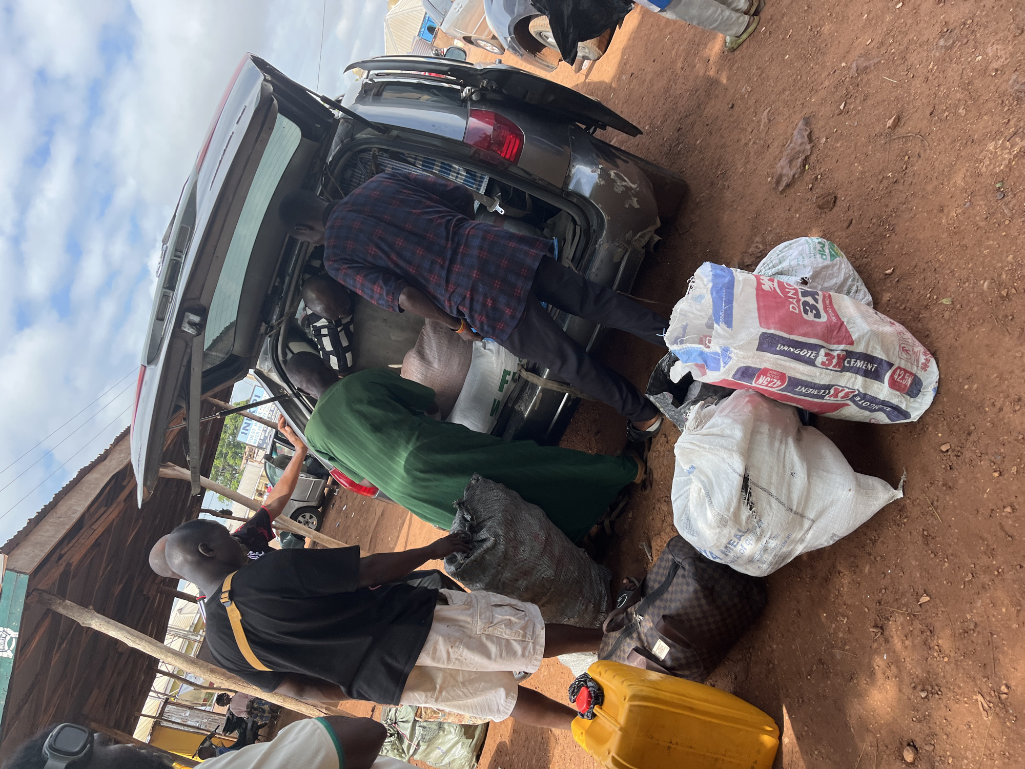The image captures an active loading scene in an outdoor, dusty environment within the city of Idah, Nigeria. A dark gray SUV, likely a minivan or crossover, is positioned with its tailgate fully open, revealing a heavily packed cargo area.
Three adult males are primarily engaged in loading or organizing items in the vehicle's rear.
* On the left, a man with a shaved head, wearing a black t-shirt and white cargo shorts, holds a large, grey, possibly net-like sack. His back is to the viewer, and he appears to be placing or adjusting the sack near the car.
* In the center, a man in a long, flowing green garment bends over, reaching deep into the SUV's trunk.
* To the right of him, another man, wearing a red and blue plaid short-sleeved shirt and dark trousers, is also bent over, actively arranging items within the vehicle's packed interior.
The SUV's rear bumper shows visible scuffs and wear. Inside the open trunk, numerous large, irregularly shaped sacks and bundles are crammed, indicating a substantial load.
On the reddish-brown dirt ground in the immediate foreground and surrounding the vehicle, several items are visible:
* A bright yellow plastic jerrycan, possibly for fuel or water, with a red cap/accessory on top.
* A dark brown duffel bag or travel bag featuring a checkered pattern.
* Multiple large white woven sacks. One prominently displays "DANGOTE 3X CEMENT 42.5KG PORTLAND CEMENT" in red, white, and blue text. Another white sack has partial text visible, including "HEALTH."
In the background to the left, a rudimentary structure with a corrugated metal roof is supported by rough wooden poles. A partially visible sign above it reads "MURITI." Further left, a person is seated on a bench next to a parked motorcycle. On the far right, part of another silver car is visible. The sky above is partly cloudy with scattered white cumulus clouds, and overhead utility lines are discernable. The ground is consistently a reddish-brown earth with some small stones and debris.