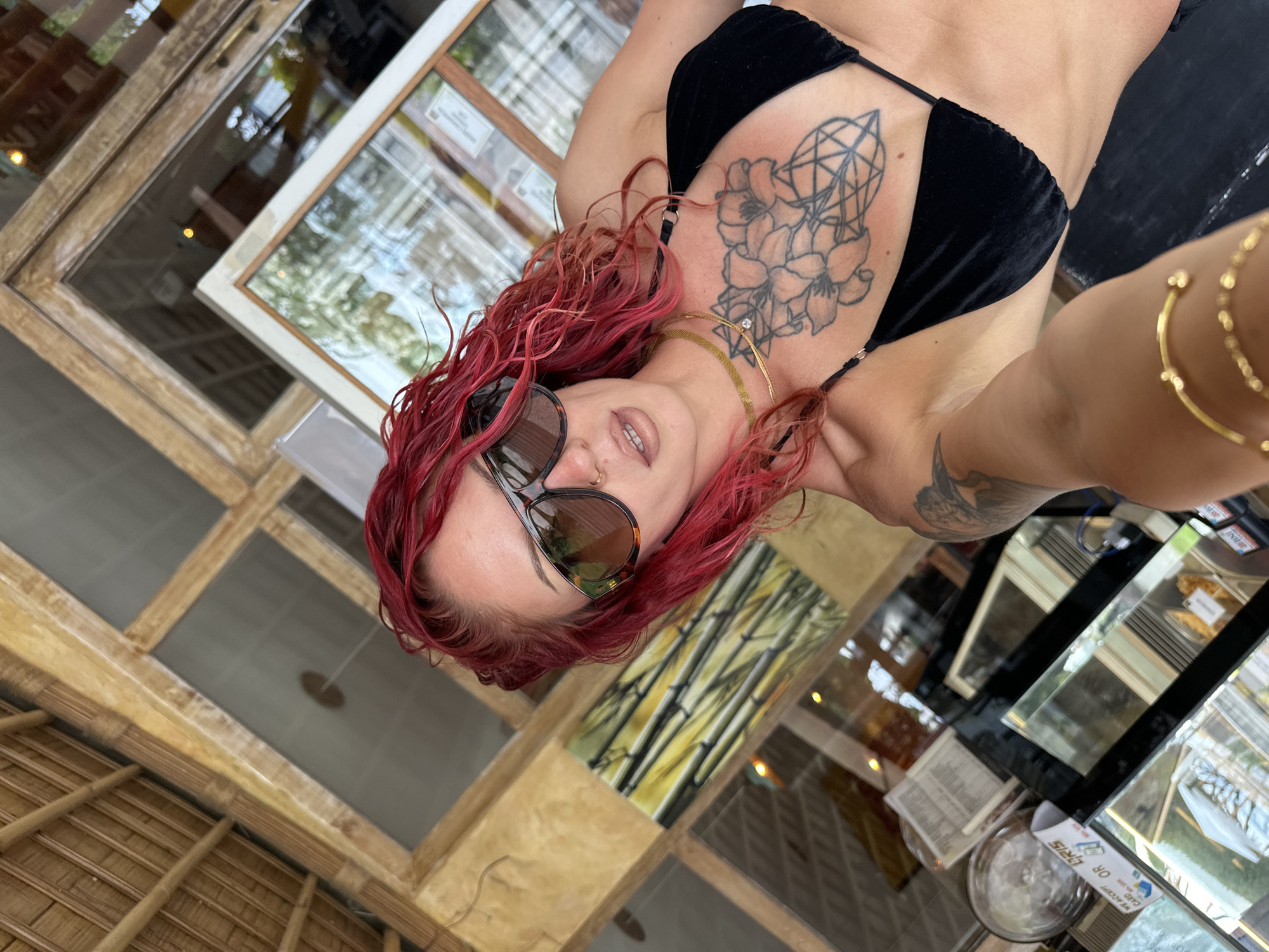 A woman with red hair and sunglasses is taking a selfie in an indoor location in Unknown, Indonesia. She is wearing a black bikini top and gold jewelry, including a necklace and bracelets. She has tattoos on her chest and arm. The background includes a window with a view of trees, a bamboo ceiling, and a display case.