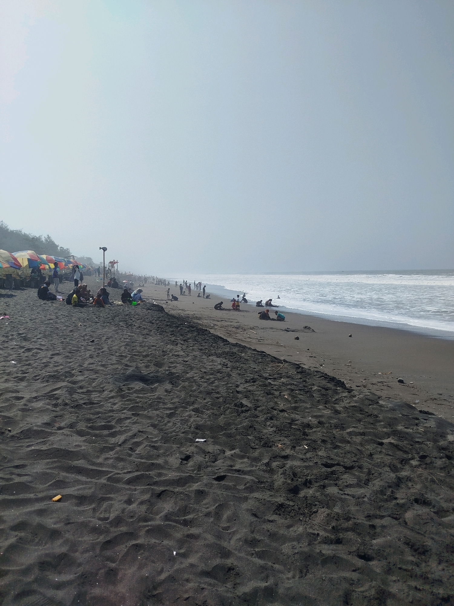 This is a wide shot of a crowded black sand beach in Kenoyajaya, Indonesia. In the foreground, the dark sand is marked with footprints and debris. In the midground, several groups of people are seated on the sand, some under colorful umbrellas. There are also people standing and walking along the shoreline. In the background, the ocean meets the sky in a hazy expanse, with waves breaking and creating white foam. The overall atmosphere is that of a sunny, busy beach day.