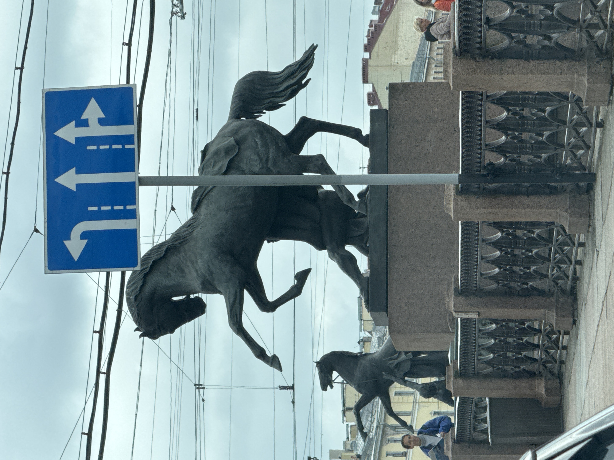 This image depicts a street scene in Saint Petersburg, Russia, featuring a large bronze equestrian statue on a granite pedestal. The statue appears to be part of the Anichkov Bridge, famous for its horse sculptures. In the background, there are buildings with typical Saint Petersburg architecture, including mansard roofs. Overhead, a complex network of power lines and tram wires crisscrosses the sky, which is overcast. A blue traffic sign with white arrows indicating lane usage is prominently visible in the upper right quadrant of the image. In the foreground, a decorative metal fence runs along the embankment. Several individuals are visible: a person in a blue jacket stands near the fence on the left, looking towards the viewer and holding some papers. Further to the right, two women are standing near the fence, one in a light-colored jacket and the other in an orange jacket. The lower left corner of the image shows the edge of a black vehicle, suggesting the photograph was taken from a moving car. The overall atmosphere is urban and somewhat somber due to the cloudy weather.