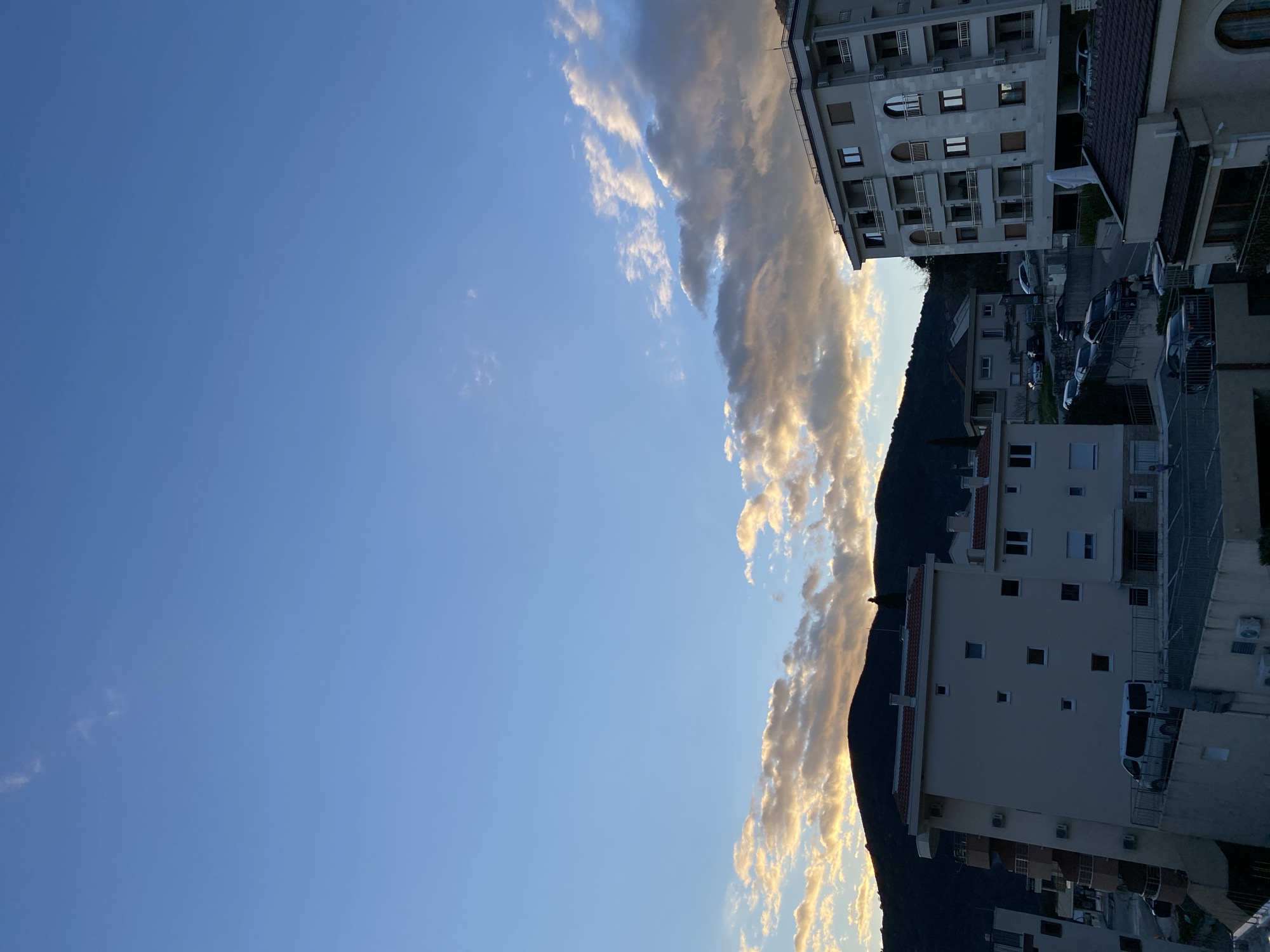 A photograph shows a vertical view of Budva, Montenegro at sunset. The clear blue sky is punctuated by a line of striated, white and gold-tinged clouds. Buildings including residential complexes, parking areas with parked cars, and a dark mountain are visible. No people are observed in the scene.