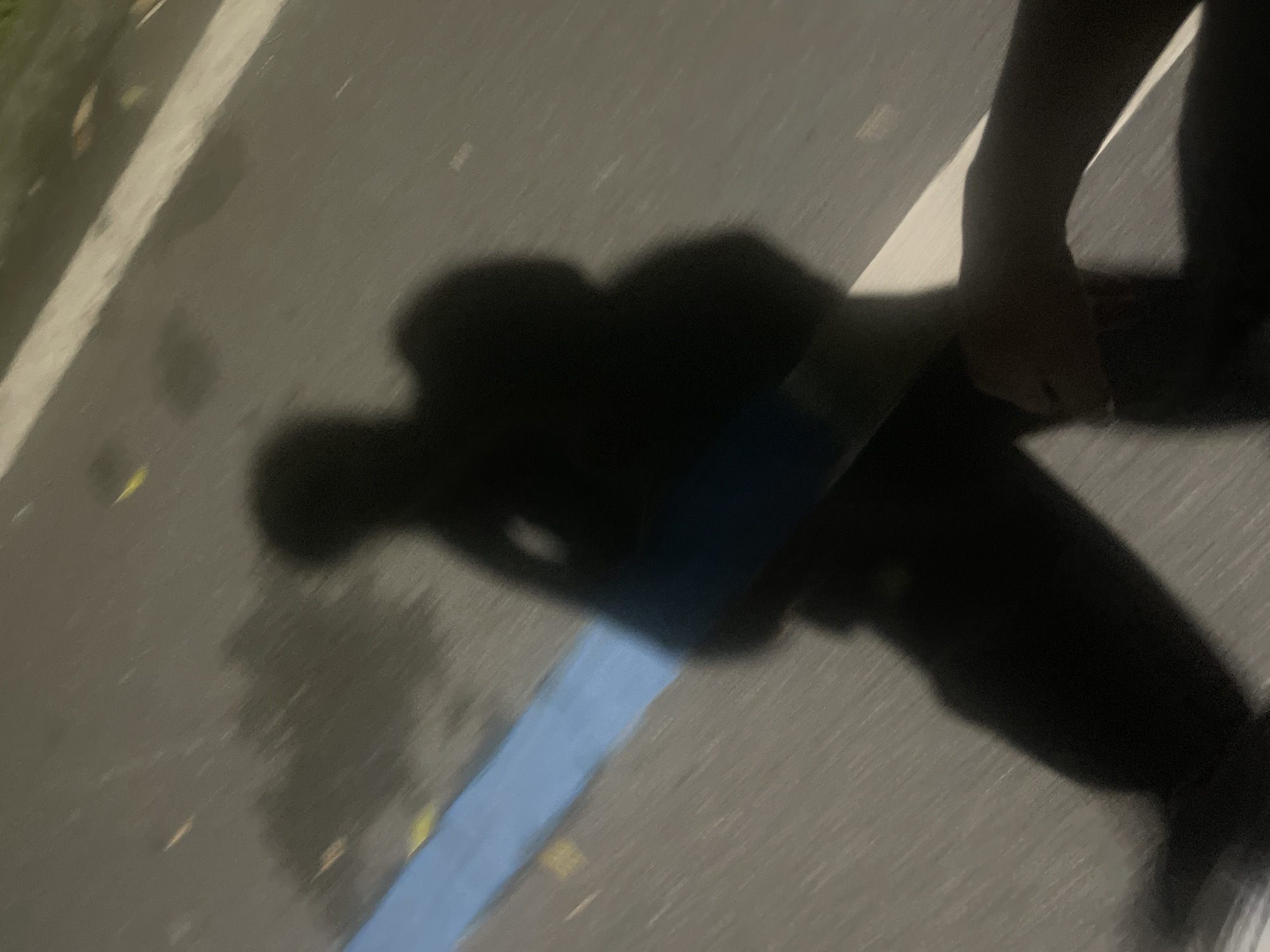 A photograph taken at night, presumably on a street or sidewalk in the Chongqing High-tech Zone, China, shows a dark, blurry shadow of a person holding a rectangular object. The background is a light-colored paved surface with a blue stripe. The shadow extends diagonally across the frame. The shadows suggests a person standing with something in their hand. The surrounding surface shows signs of dampness or recent rain.