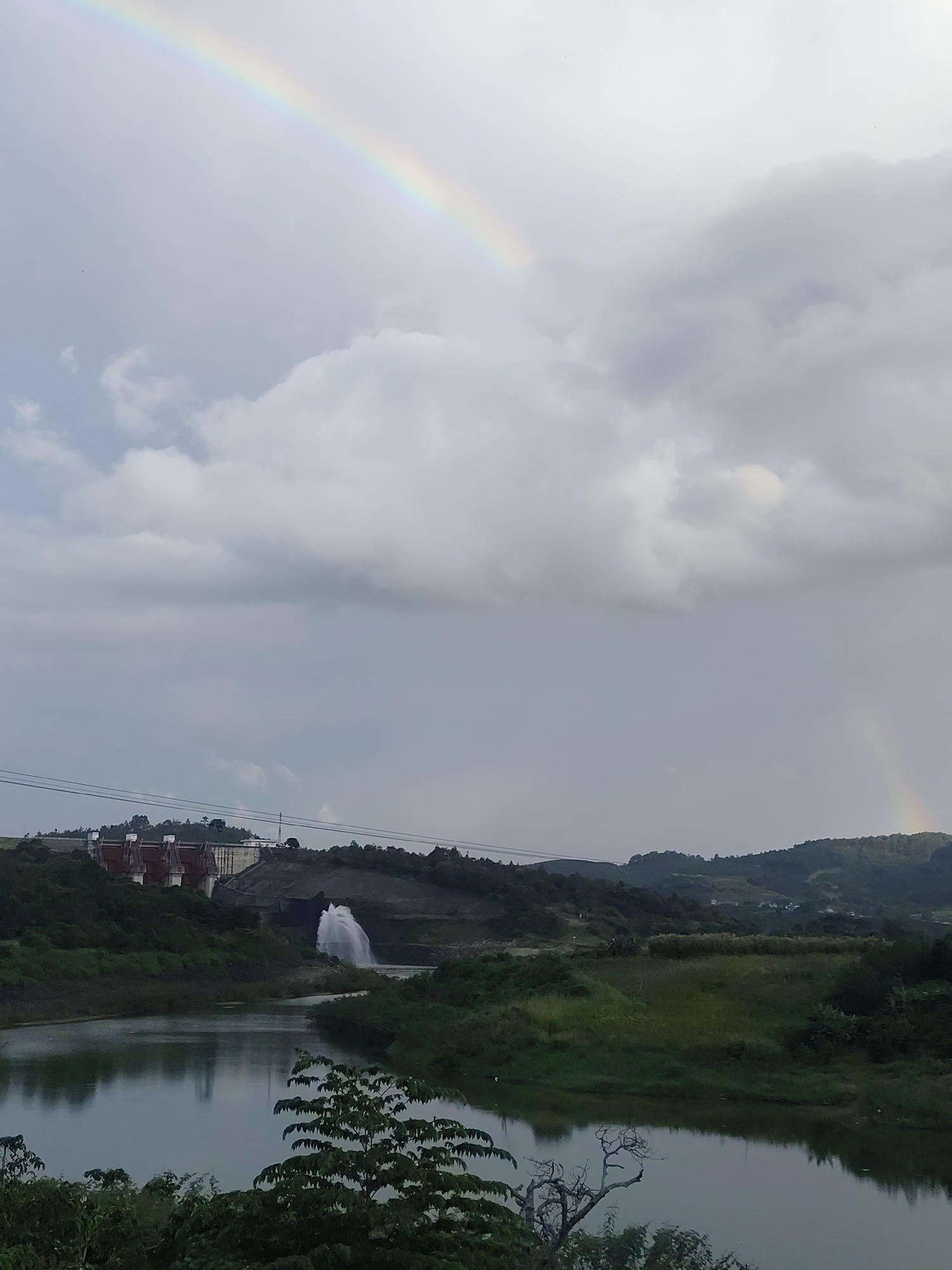 The image shows a wide shot of a landscape with a dam and a body of water in the foreground. The sky is overcast with clouds, and a rainbow is visible in the upper left quadrant of the image. The dam, a large concrete structure with multiple towers, is situated on a hillside in the middle ground. Water is being discharged from the dam, creating a plume of spray. The surrounding landscape is covered in lush green vegetation. The body of water in the foreground reflects the sky and the surrounding greenery. The image captures a serene and natural environment, possibly a reservoir or a lake near the dam. The location is stated to be in the city of Phú An, Vietnam. There are no people visible in the image.