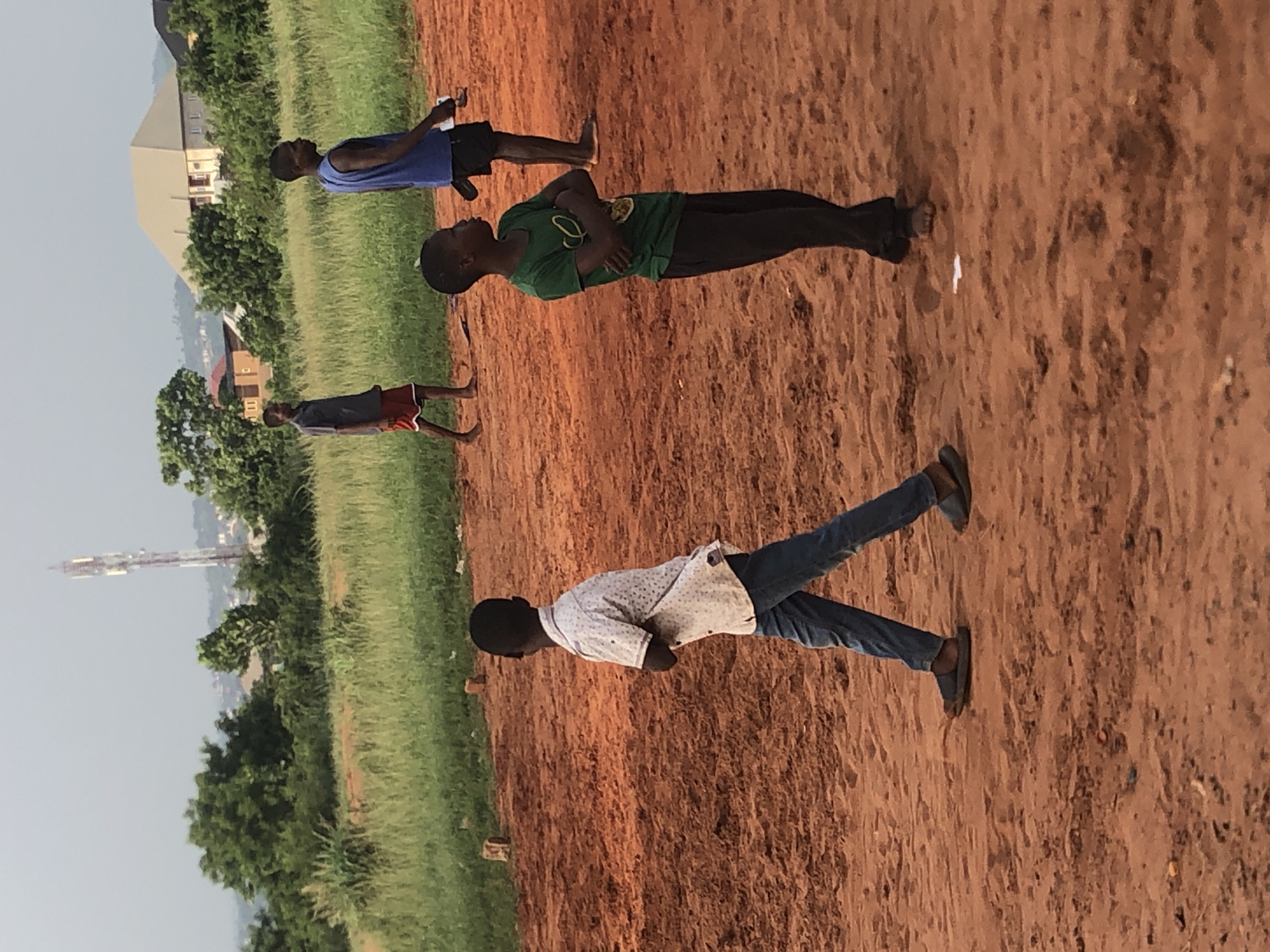 The image shows four young males standing outdoors on a reddish-brown dirt surface in what appears to be an open, unpaved area. The setting is likely in Nsugbe, Nigeria, during the daytime under a clear or slightly hazy sky.
From left to right (if rotated correctly):
One young male is dressed in a dark t-shirt and red-colored shorts, standing barefoot.
Next, another young male wears a blue tank top and dark shorts or pants, also barefoot. He appears to be holding a small white object in his right hand.
In the foreground, a young male is seen wearing a green t-shirt and dark pants, standing with his arms crossed. He is barefoot.
Further to the right and slightly closer to the viewer, another young male wears a light-colored, possibly patterned, short-sleeved shirt and blue jeans. He stands with his arms crossed and is wearing dark sandals.
Behind the individuals, there is a band of green grass and trees, separating the immediate dirt area from the distant background. In the background, various buildings are visible, including a prominent light-colored structure with a distinctive roof. A tall, slender tower or mast can also be seen rising in the far distance against the sky. The scene depicts a casual gathering with no specific active event taking place, but rather a relaxed posture among the youths. No visible text is present in the image.