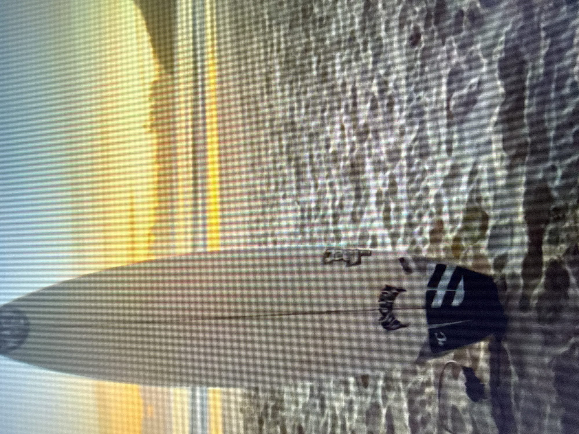 A surfboard stands upright on a beach with sand visible in the foreground and water and a sunset in the background. The surfboard is white with a black logo at the top that appears to read "WET" in a circle. Below that, there is another black logo that reads "LOST". The tail of the surfboard has black and white stripes. A leash is attached to the tail. The sunset is casting a warm glow over the water and the sky, with hues of orange, yellow, and blue. The water appears calm with gentle waves. The beach is covered in sand with some ripples and shadows indicating texture. The scene is calm and serene.