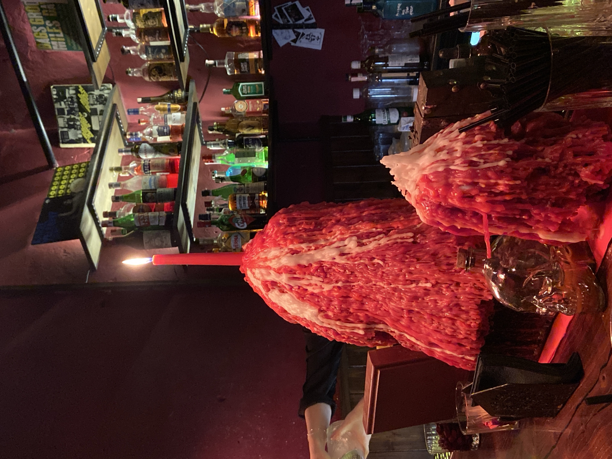 The image depicts a dimly lit bar scene, likely in Brest, Belarus, given the context. In the foreground, two large, dripping red candles with white wax are prominently displayed. The candle on the left is taller and more conical, while the one on the right is shorter and wider. A single red candle with a lit flame stands atop the left candle. To the left of the candles, a glass containing what appears to be red berries is visible, next to an empty glass. A dark, angular object, possibly a coaster or holder, sits between the glasses and the large candles. Further to the right, a glass bottle shaped like a human skull, containing a golden liquid, rests on the wooden bar surface. Behind the candles, a wooden bar counter is visible, cluttered with various items. Shelves in the background are stocked with numerous bottles of alcohol, illuminated by a warm, ambient light. Some framed pictures and posters are affixed to the wall above the shelves. The overall atmosphere suggests a unique and possibly gothic-themed bar.