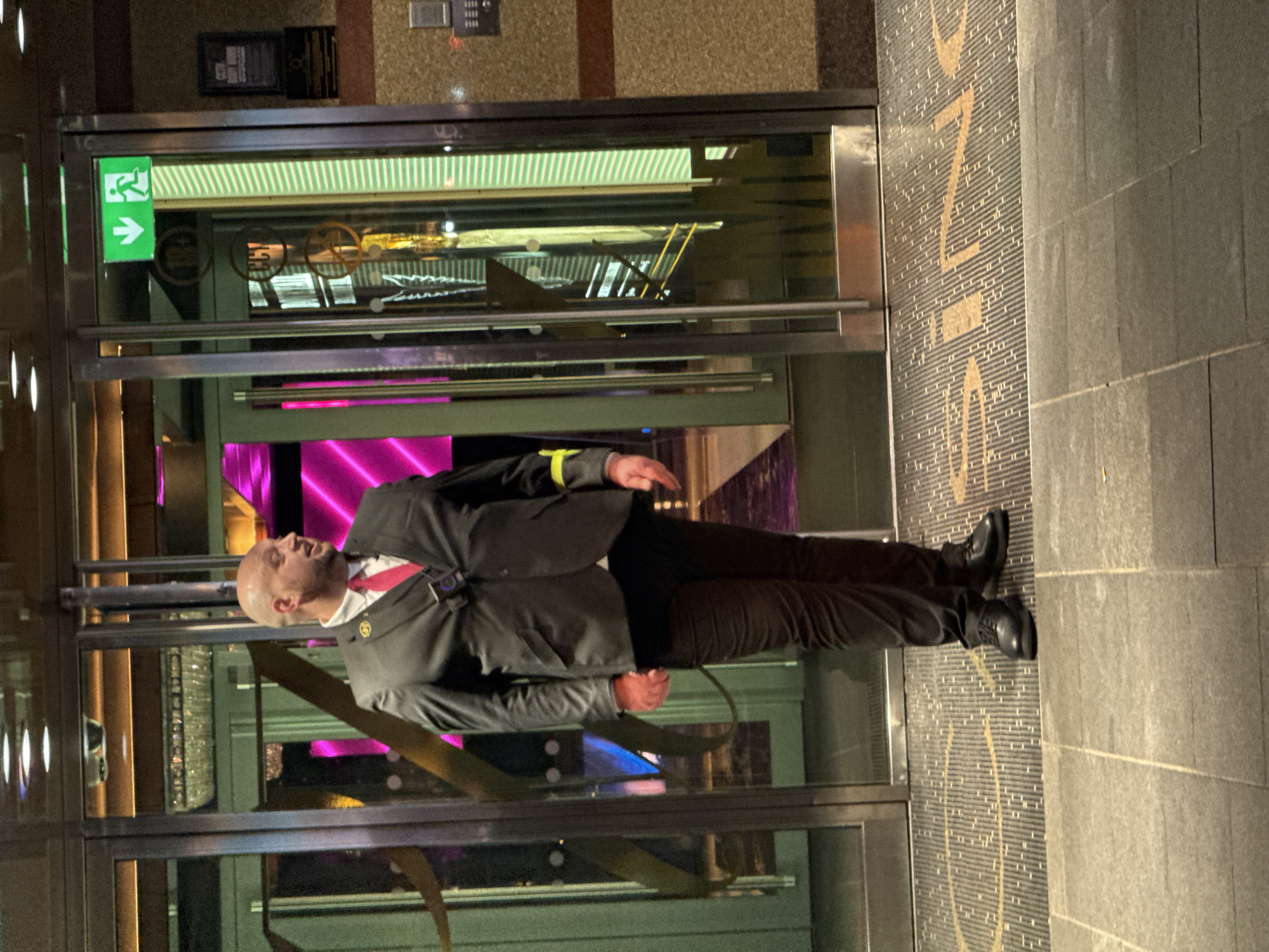 A man in a suit stands at the entrance of what appears to be a hotel or event venue in London, United Kingdom. The man, who is bald, wears a black suit jacket with a yellow armband, a white shirt, and a red tie. He also has a security badge. The entrance features glass doors with an exit sign and gold lettering on the exterior. Inside, the venue is brightly lit with a mix of green, gold, and pink lighting. The exterior ground surface is a patterned stone.