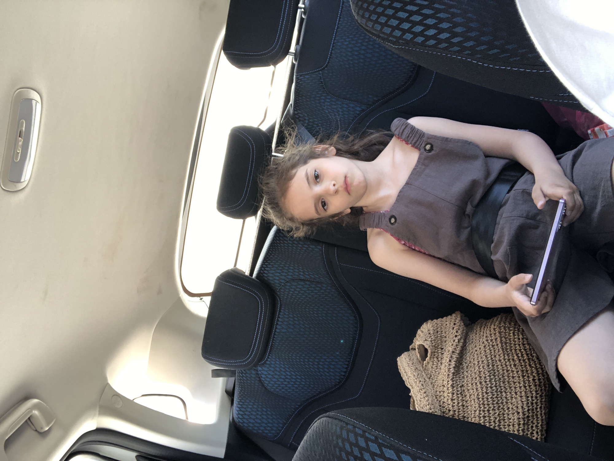 The image presents the interior of a vehicle, specifically the rear passenger compartment, during daylight hours. A young girl, appearing to be approximately 8-10 years old, is seated in the rear passenger seat, directly facing the camera with a neutral expression. She has light brown, curly hair and is wearing a dark brown or charcoal-colored sleeveless romper with button details on the shoulder straps and a black belt. She holds a light purple smartphone horizontally with both hands.
The car's interior features dark grey or black fabric seats with a distinctive blue geometric diamond-like pattern on the backrest and contrasting light blue stitching outlining the seat and headrest contours. Three headrests are visible across the rear bench, one partially obscured by the girl. On the seat to the girl's left, a large, light brown, intricately woven bag, possibly made of straw or jute, rests. The vehicle's ceiling is light beige, exhibiting some minor stains or smudges. An oblong overhead light fixture with a switch is centrally mounted on the ceiling, and a light-colored grab handle is visible on the upper left side near a window frame. Bright natural light illuminates the cabin, entering through the rear side windows. The specific location within the city of Saint Petersburg, Russia, cannot be determined from the visual information present in the image.