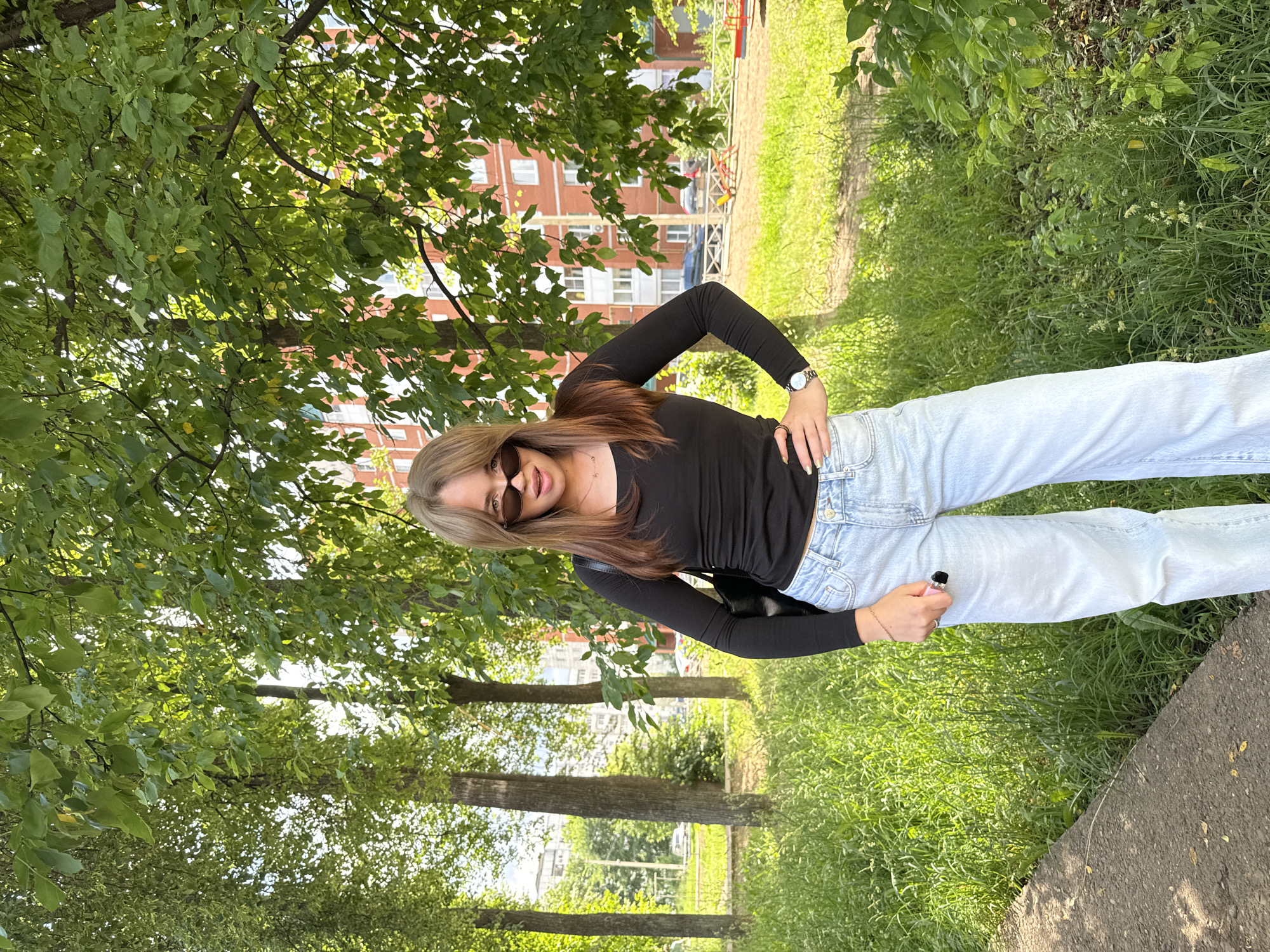 A young woman wearing sunglasses, a black long-sleeved shirt, and light blue wide-leg jeans stands outdoors in Chaikovsky, Russia. She is positioned in the foreground, with a street, residential buildings, and trees in the background. The woman has her left hand on her hip and is holding a pink vape device in her right hand. The environment appears to be a park or green space, with lush grass and trees providing shade. The sun is shining, casting shadows and illuminating the scene.