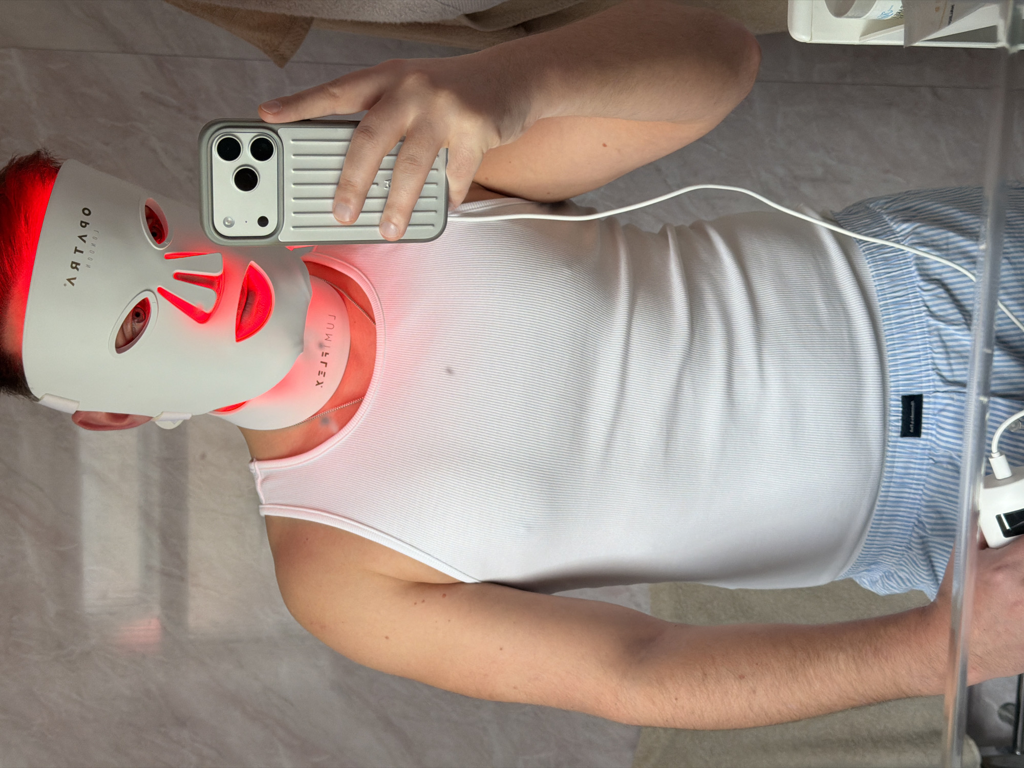 A person is lying on their back, wearing a white tank top and blue striped pajama pants. They are also wearing a white LED facial mask with red lights. The mask obscures most of their face, but their eyes are visible through the eye openings. The mask is connected to a white cord. The person is holding a smartphone in their right hand, taking a selfie or a photograph. The background appears to be a bathroom, with light-colored tile flooring. The text "London" is visible on the mask.