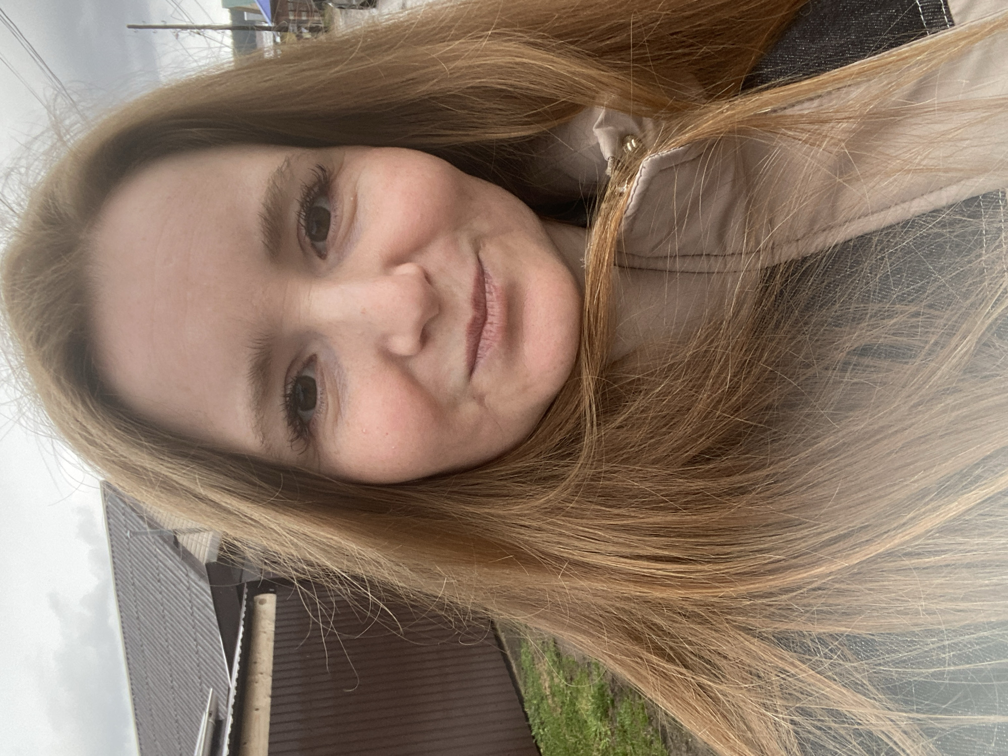 A close-up, off-center, rotated, and seemingly hand-held selfie shows a fair-skinned woman with long, straight, light brown-blonde hair. She is smiling slightly. She is wearing what appears to be a light-colored jacket with a denim collar. In the blurry background is a section of a building with a brown exterior, green foliage, and utility poles with wires. Based on the available information, the location is identified as Chaikovsky, Russia.