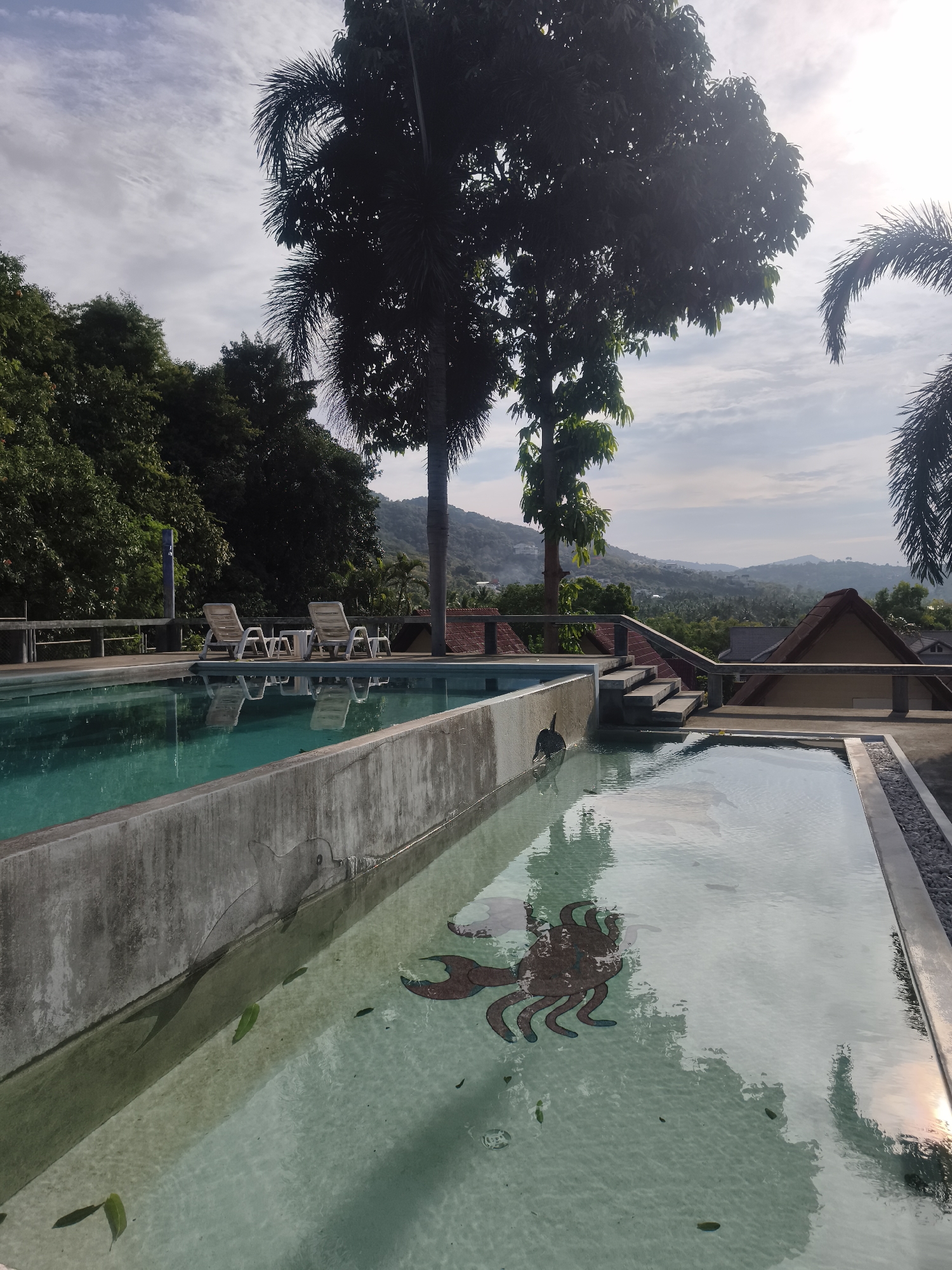 The image shows a two-level swimming pool complex in บ้านบางรักษ์, Thailand. The upper pool, filled with turquoise water, features two sets of lounge chairs on its deck. Adjacent to it is a lower, elongated pool filled with lighter-colored water. A mosaic crab and dolphin are visible at the bottom of the lower pool. Steps connect the two pools. Lush tropical vegetation surrounds the complex, with mountains visible in the background. The sky is partly cloudy.