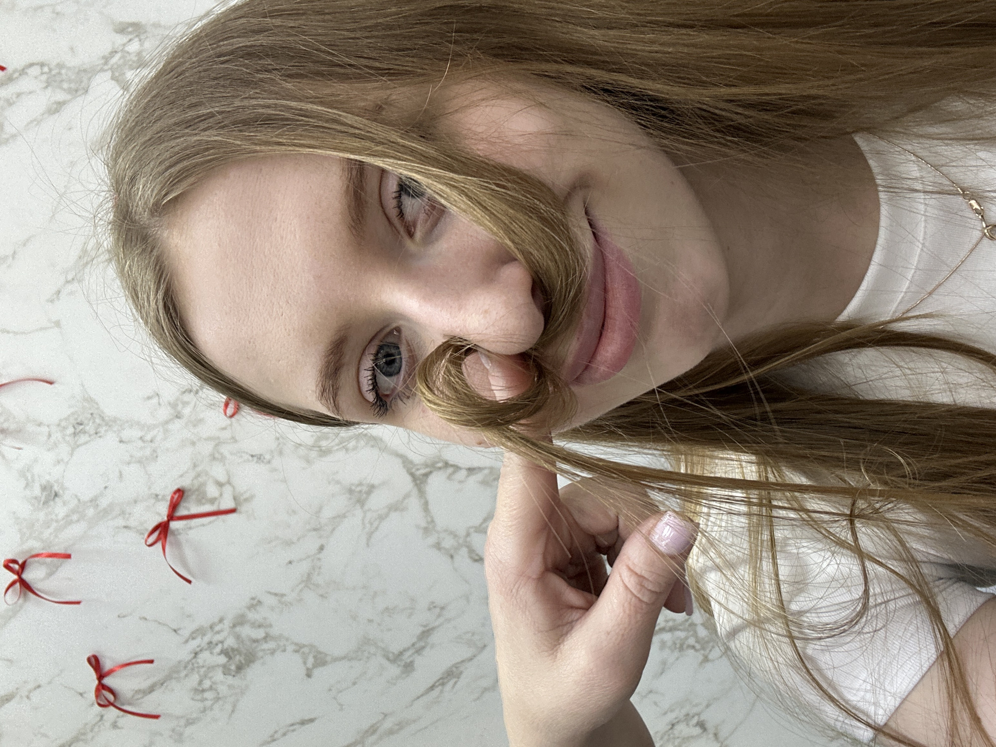 A fair-skinned young woman with long, light-brown hair holds a strand of her hair under her nose, creating the illusion of a mustache. She is wearing a white t-shirt and a thin gold necklace. Her nails are painted light pink. The background is a faux marble surface with scattered small red ribbons. No information is available to identify this picture with the city of Chaikovsky, Russia.