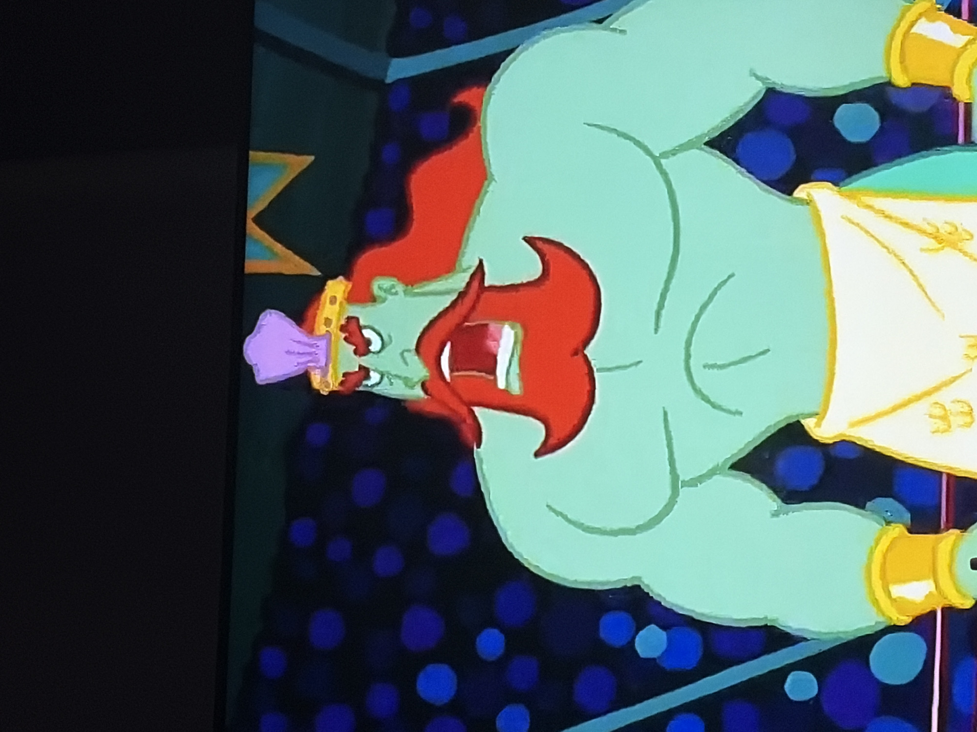 The image is a close-up of an animated character, likely from a cartoon. The character is a muscular, green-skinned male with a bright red beard and long, red hair. He is wearing a golden crown adorned with a purple seashell-like object on top. He has an angry expression with his mouth wide open, revealing his teeth and pink tongue. He is also wearing a white loincloth or skirt with gold trim. Around his wrists, he has thick gold bracelets. The background appears to be a dark space with large, blurry blue circles. There is also a hint of a teal-colored structure with a golden zigzag pattern at the top. There is no discernible connection to the city of Goryachiy Klyuch, Russia.