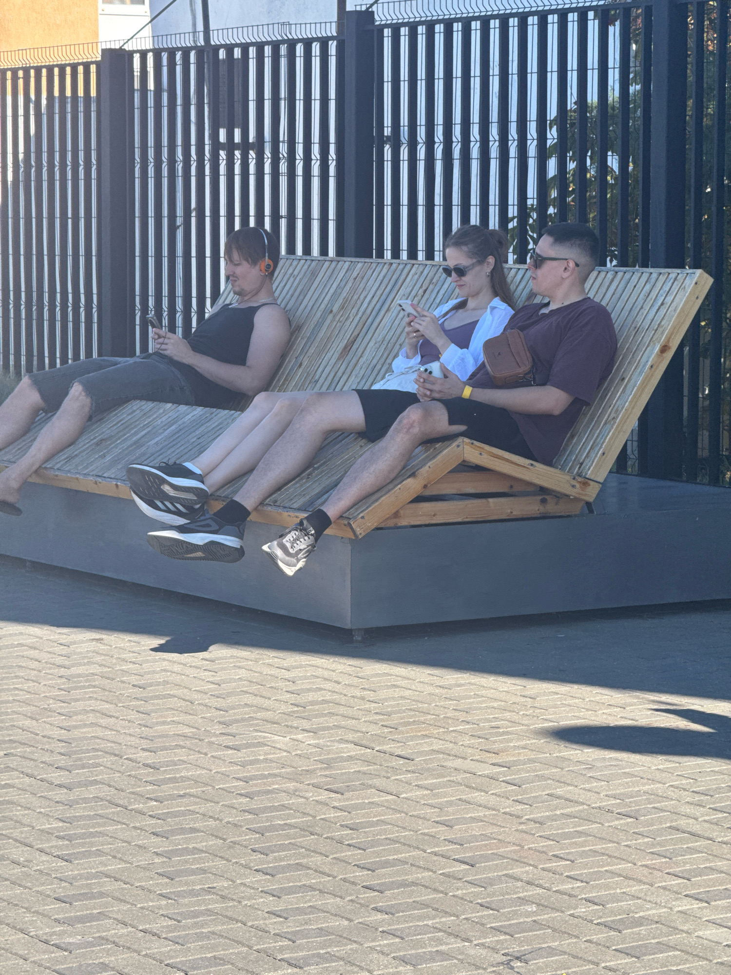 Three people are sitting on a wooden bench in Anapa, Russia. The person on the left is a man wearing a black tank top and dark shorts, with orange headphones on. He is holding a smartphone. The person in the middle is a woman wearing a white shirt over a purple top and dark shorts, with sunglasses. She is holding a smartphone. The person on the right is a man wearing a maroon t-shirt and dark shorts, with sunglasses and a brown satchel over his shoulder. He is also holding a smartphone. The bench is positioned outdoors on a paved area with a metal fence in the background. The lighting suggests it is daytime, with shadows cast on the ground.