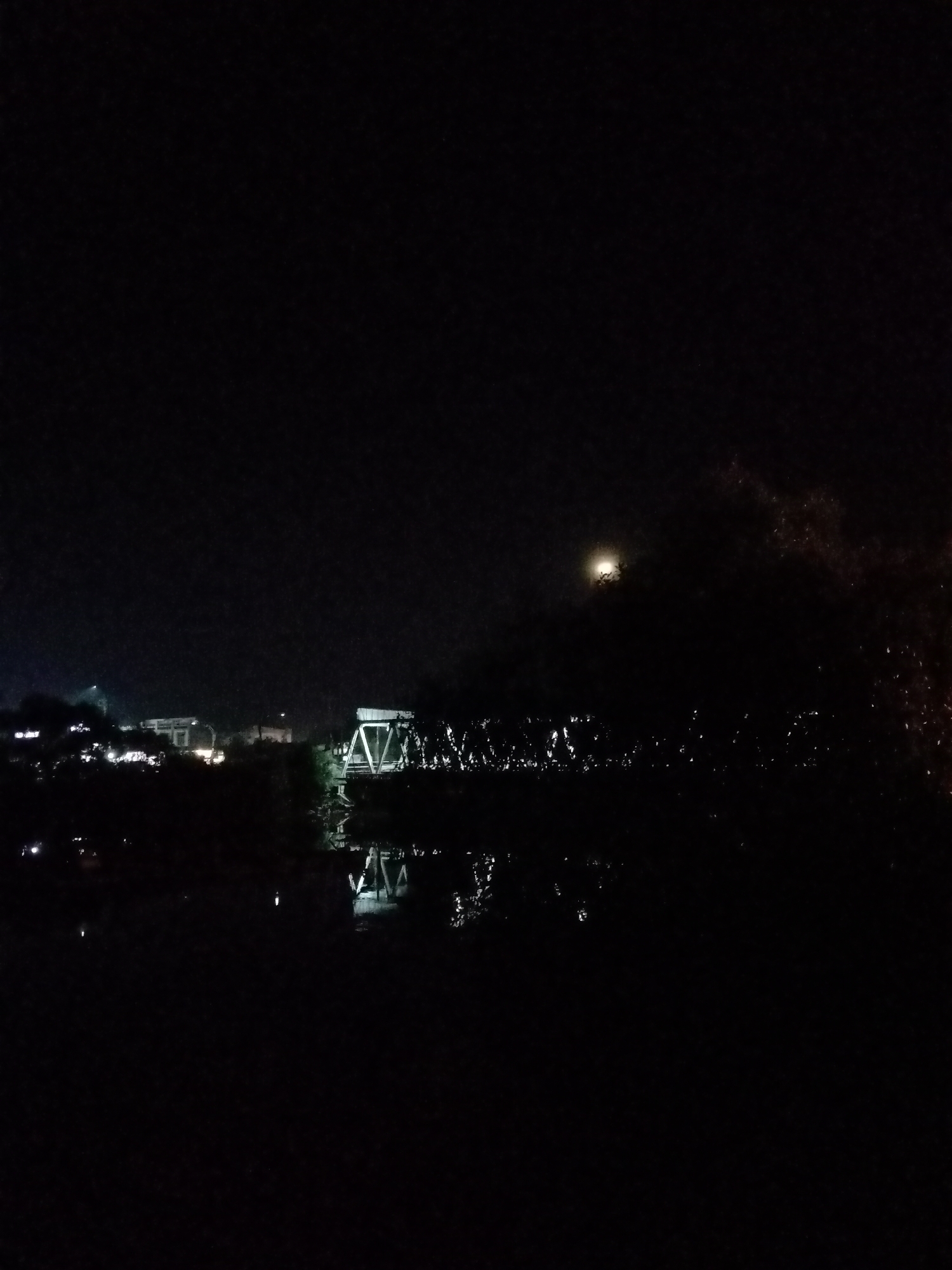 At night, the image reveals a dark scene in Lhokseumawe, Indonesia. A bridge structure with triangular supports spans a body of water, with its reflection visible on the surface. To the left, are illuminated buildings, likely part of the urban area. A bright orb, possibly the moon, is visible in the distance, partially obscured by foliage. The foreground is mostly dark, with sparse bright speckles suggesting light reflecting off of water or other surfaces.
