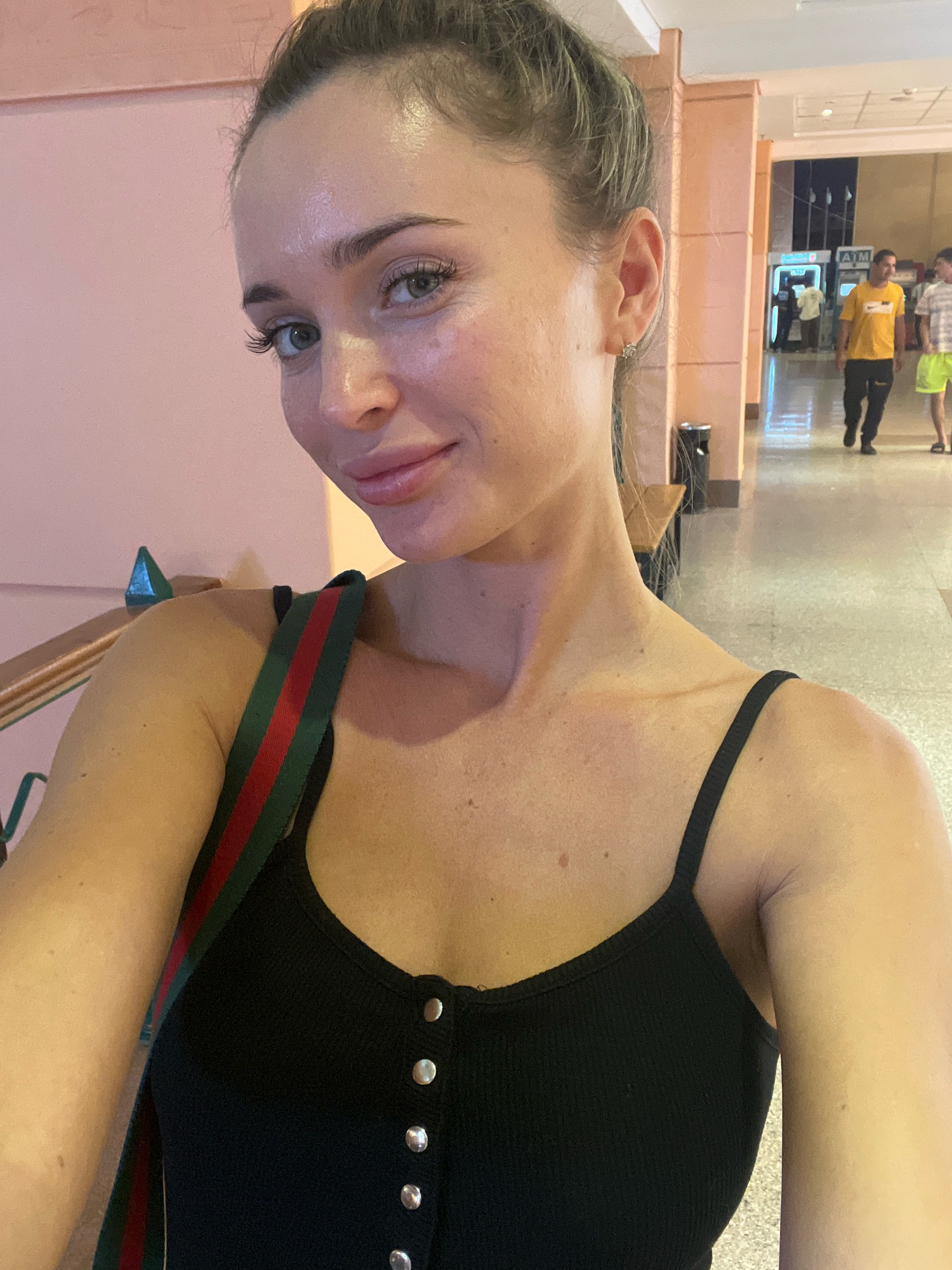 The image shows a young Caucasian woman with blonde hair pulled back in a bun, taking a selfie. She is wearing a black camisole with silver buttons. Her right shoulder is visible, and a strap from a handbag with red, green, and black stripes crosses her chest. She has light makeup on, highlighting her eyes and lips. Her expression is a slight smile.
The background suggests an indoor public space. To her right, a hallway is visible with tiled flooring and beige-colored architectural pillars. A wooden bench and a trash can are present along the hallway wall. In the distance, two men are walking. One man in a yellow t-shirt is closer to the foreground, and another man in a striped shirt and yellow shorts is further down the hall. There appears to be an ATM machine in the background. The overall lighting is bright, suggesting it could be a hotel lobby or a shopping area in a warm climate. This location is consistent with the common amenities found in hotels or resorts in Sharm El Sheikh, Egypt, which often feature such indoor common areas for guests.